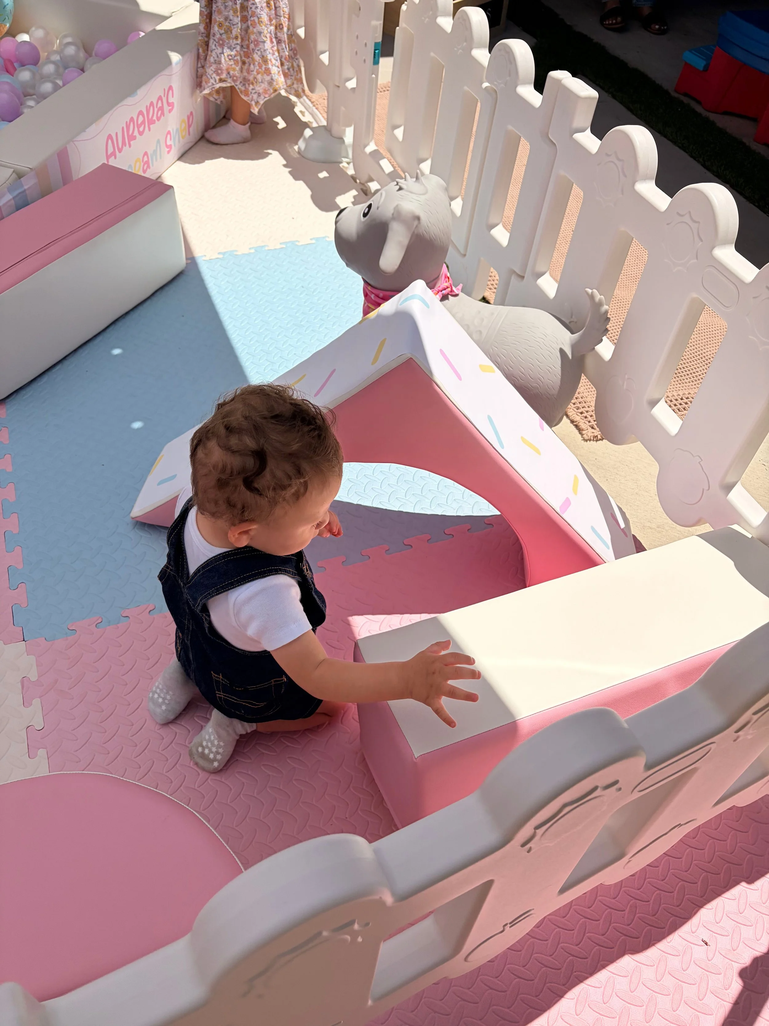 A young child kneeling on a pink foam play mat, reaching towards a pastel pink and white toy castle. The castle has a pink slide, a white fence, and a mini tunnel. A white stuffed animal resembling a bunny with a pink scarf is inside the castle, look