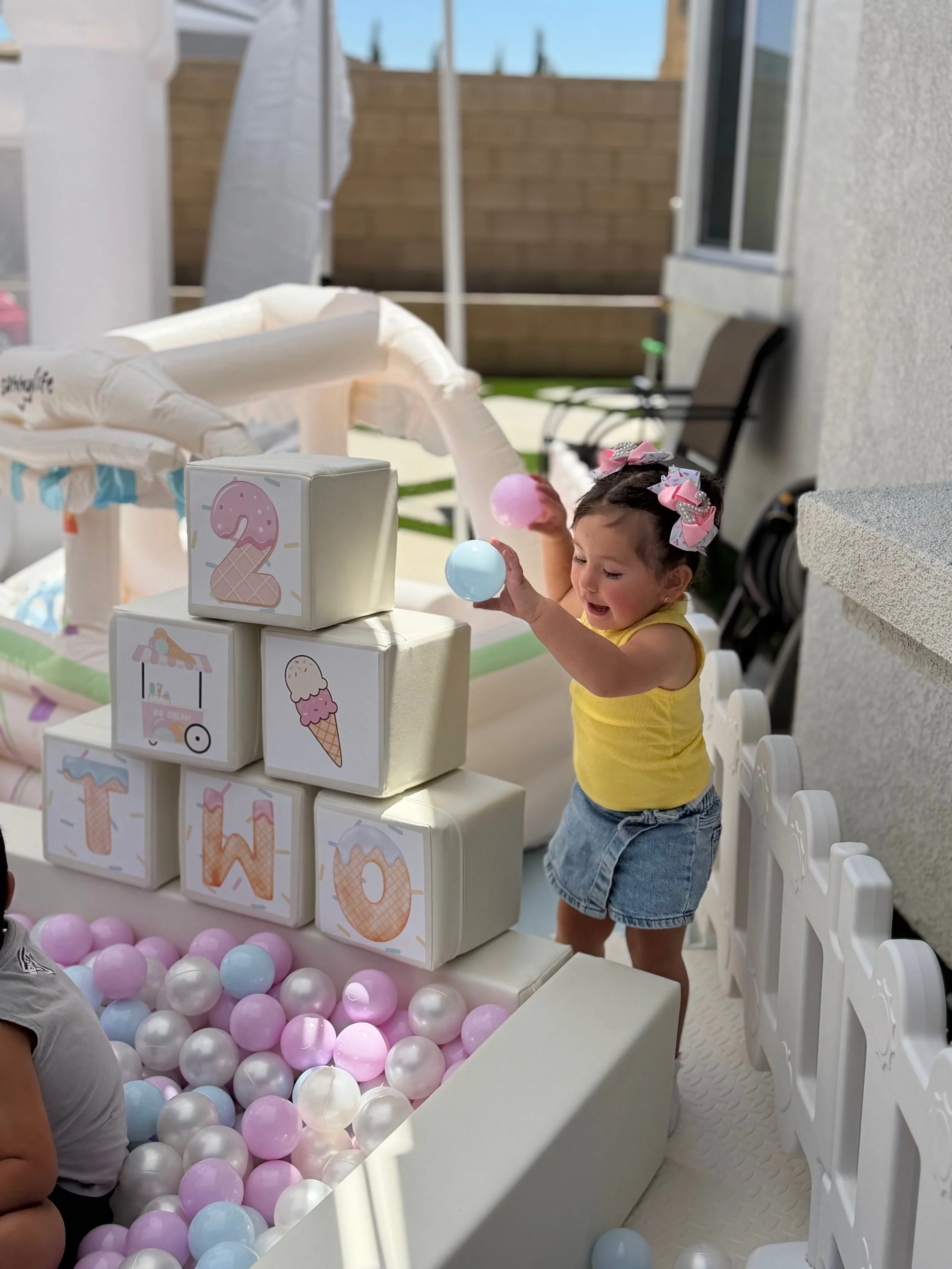 A young girl with bows in her hair playing with pastel-colored balls at a birthday party or celebration.