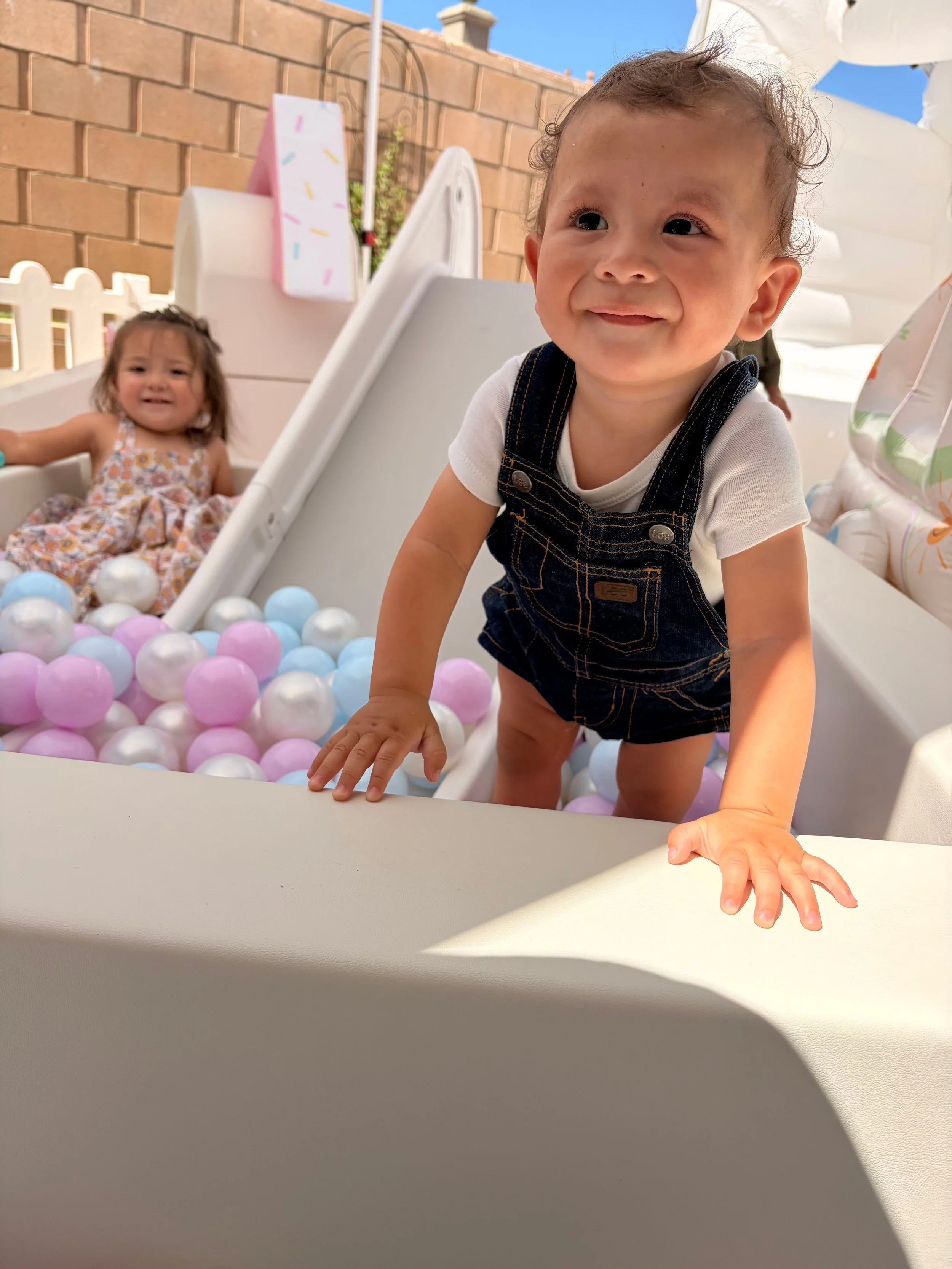A young boy in denim overalls and a white t-shirt, smiling and crawling on a ball pit with pastel-colored balls, outside under a clear sky. In the background, a girl in a floral dress sitting in the ball pit and a brick wall.