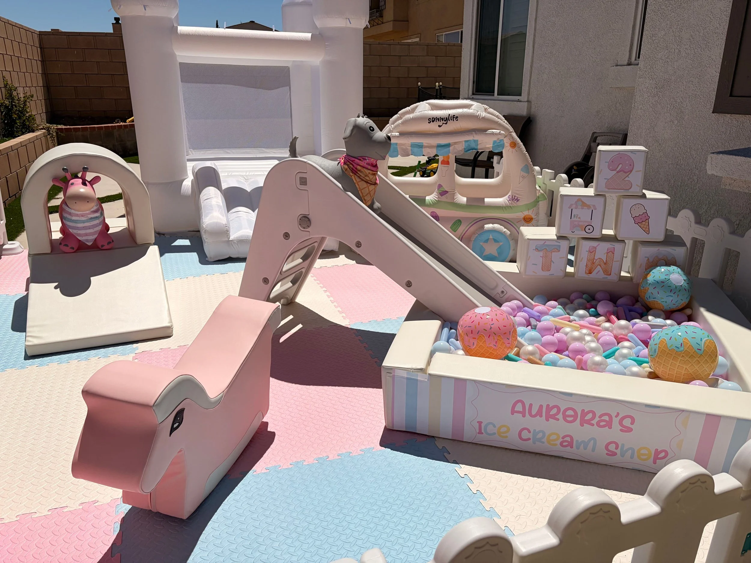 Outdoor play area with inflatable toys, pink slide, ball pit, and ice cream shop themed decor, enclosed by a white fence, with a building wall and windows in the background.