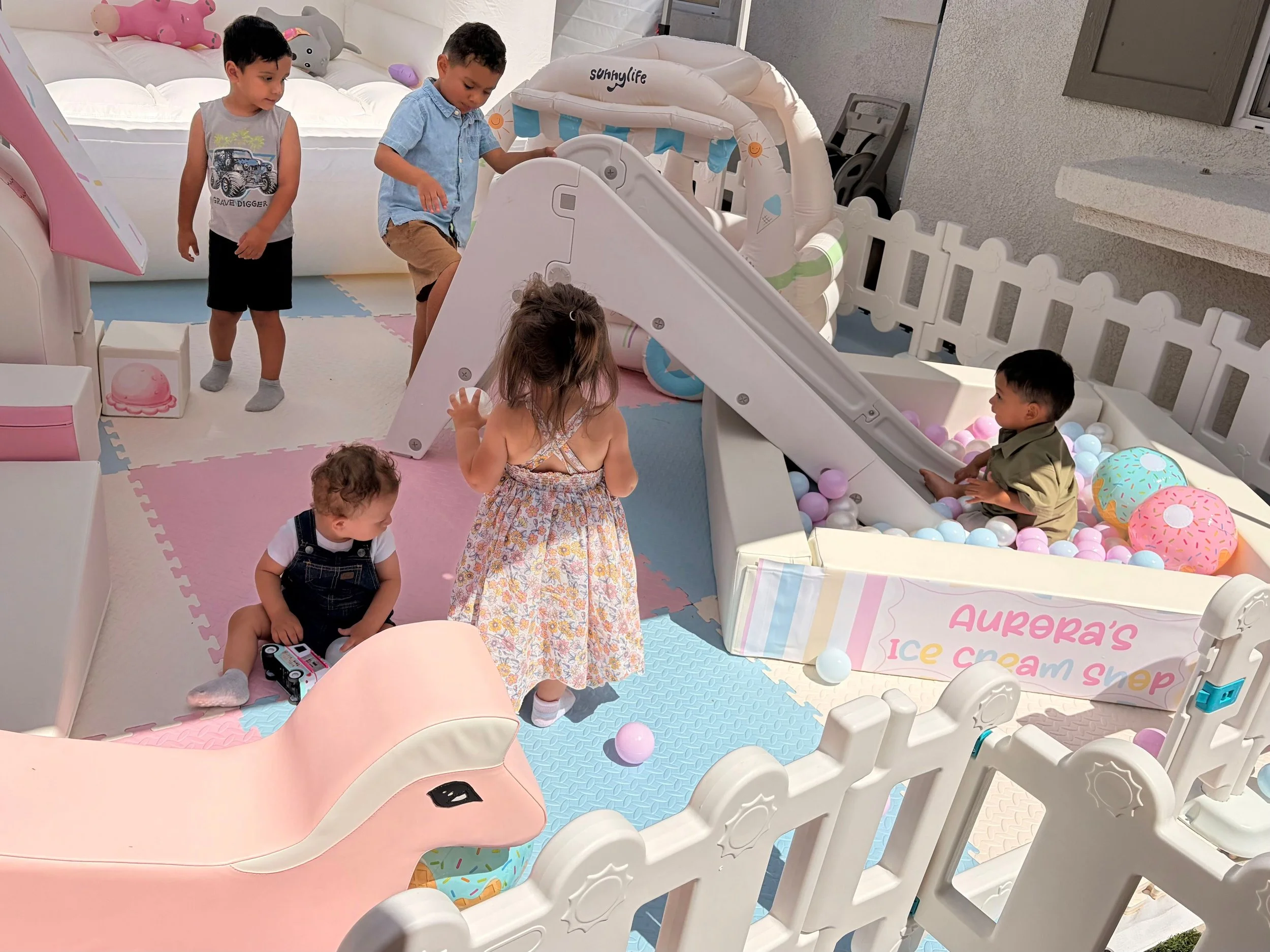 Children playing in a pastel-colored indoor play area with a small slide, ball pit, and soft foam mats, surrounded by white fencing and furniture.