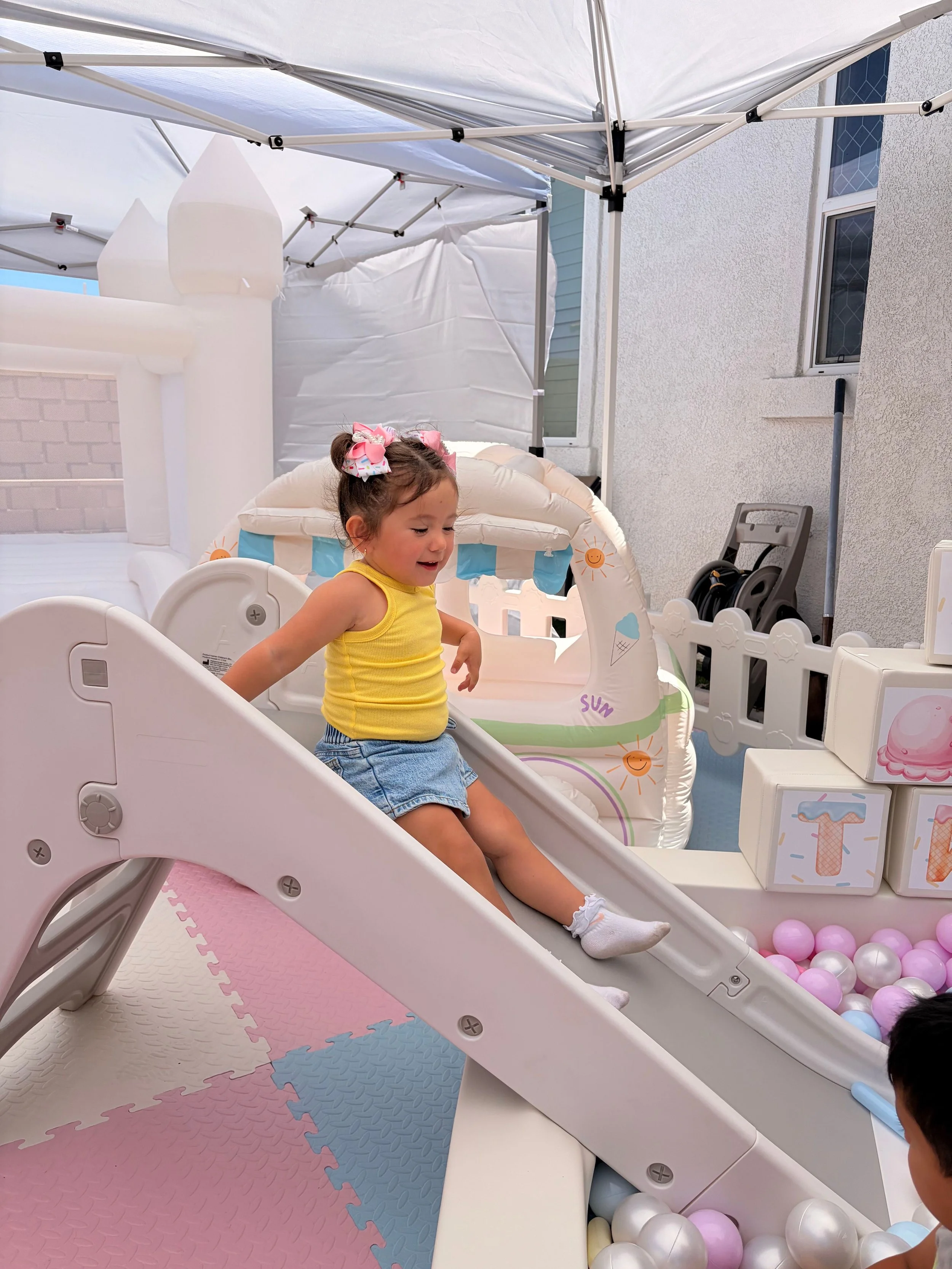 A young girl in a yellow top and denim shorts playing on a slide in a children's play area with pastel-colored foam mats, a ball pit with pink, white, and purple balls, and background elements including a white fence, a playhouse, and shade canopy.