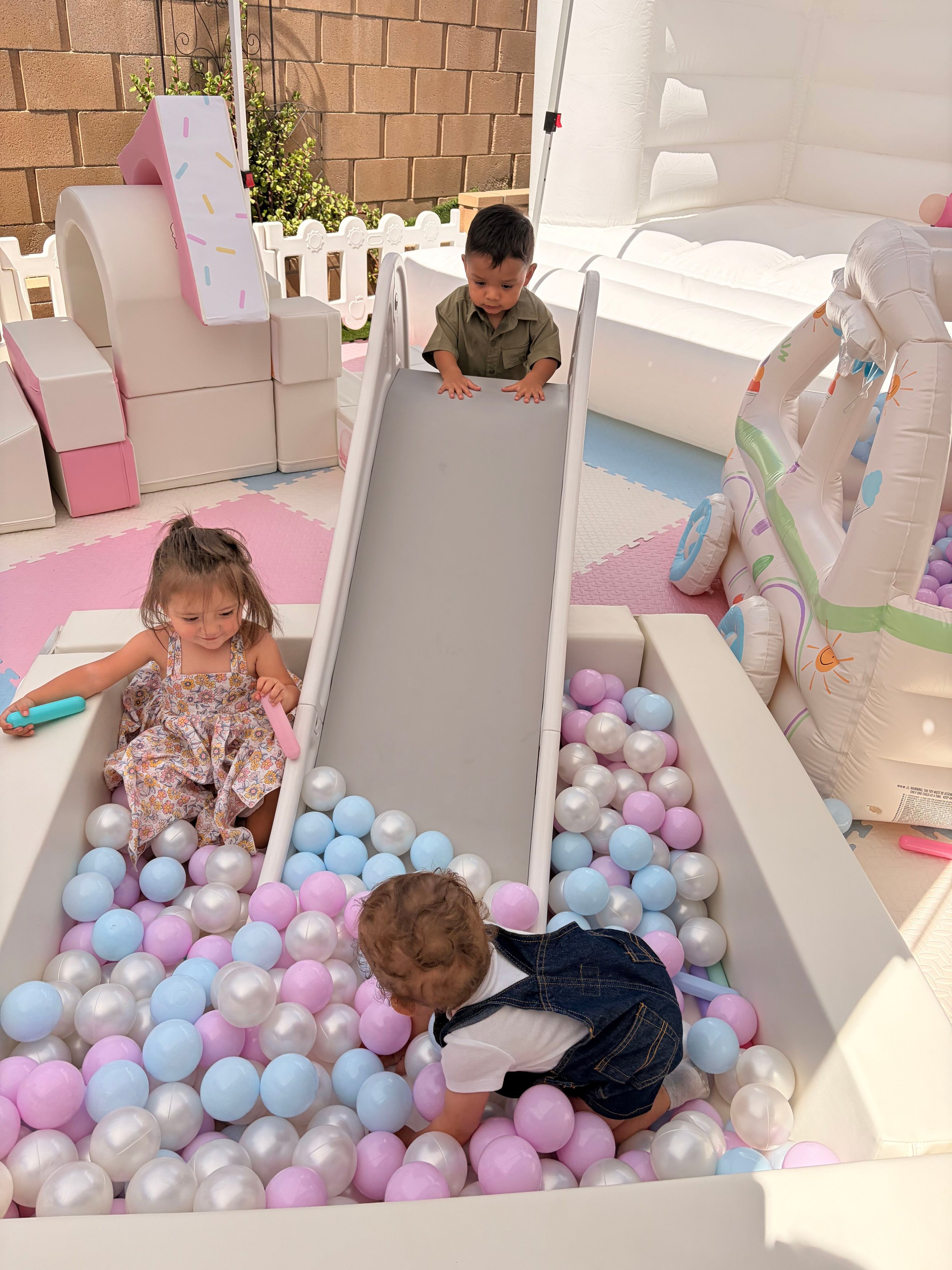 Children playing in a ball pit filled with pink, blue, and white plastic balls, with a small slide and inflatable castle in the background.