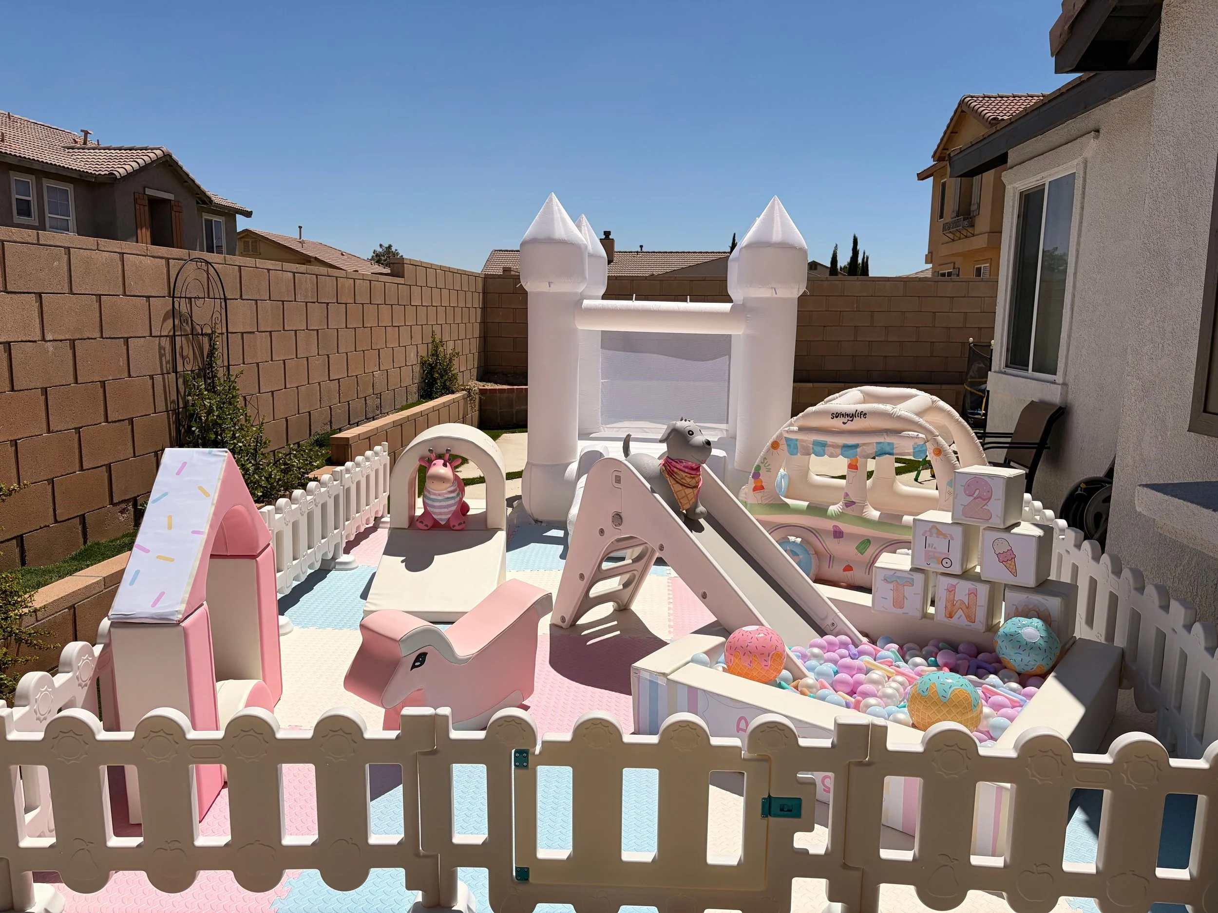 Colorful outdoor children's play area with a small inflatable castle, slide, and various toys, enclosed by a white picket fence, with a brick wall and neighboring houses in the background on a sunny day.