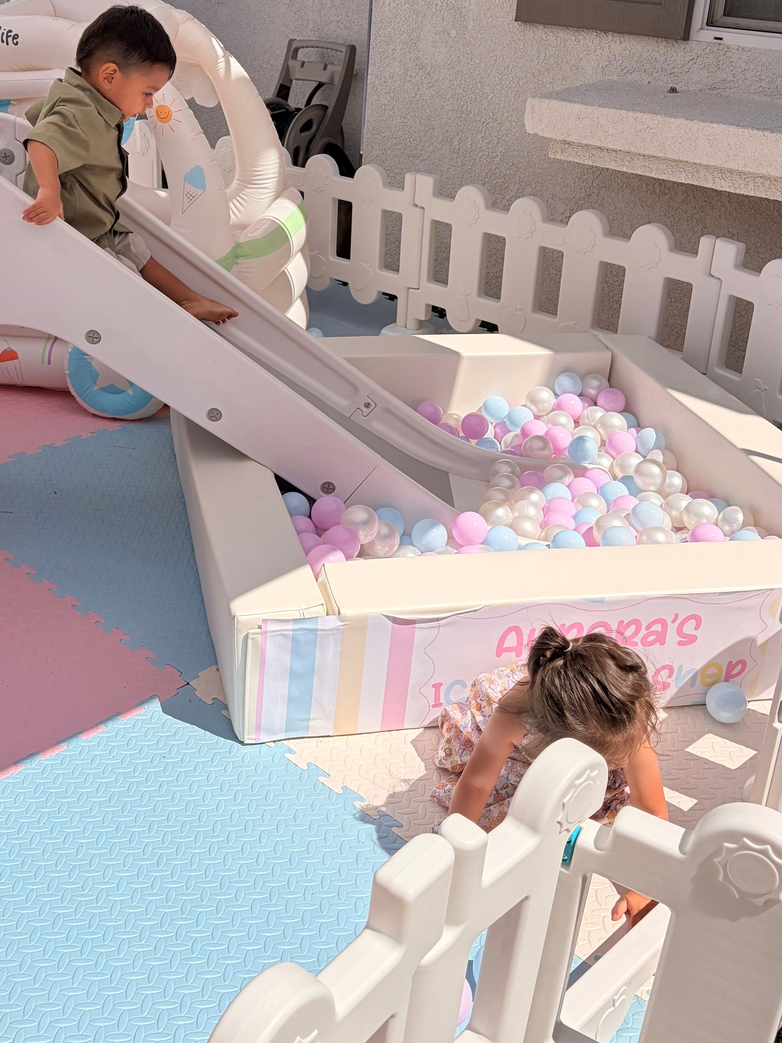 Child playing in a ball pit filled with pink, white, and blue plastic balls, surrounded by a white plastic safety fence and slide, with a gray wall and window in the background.