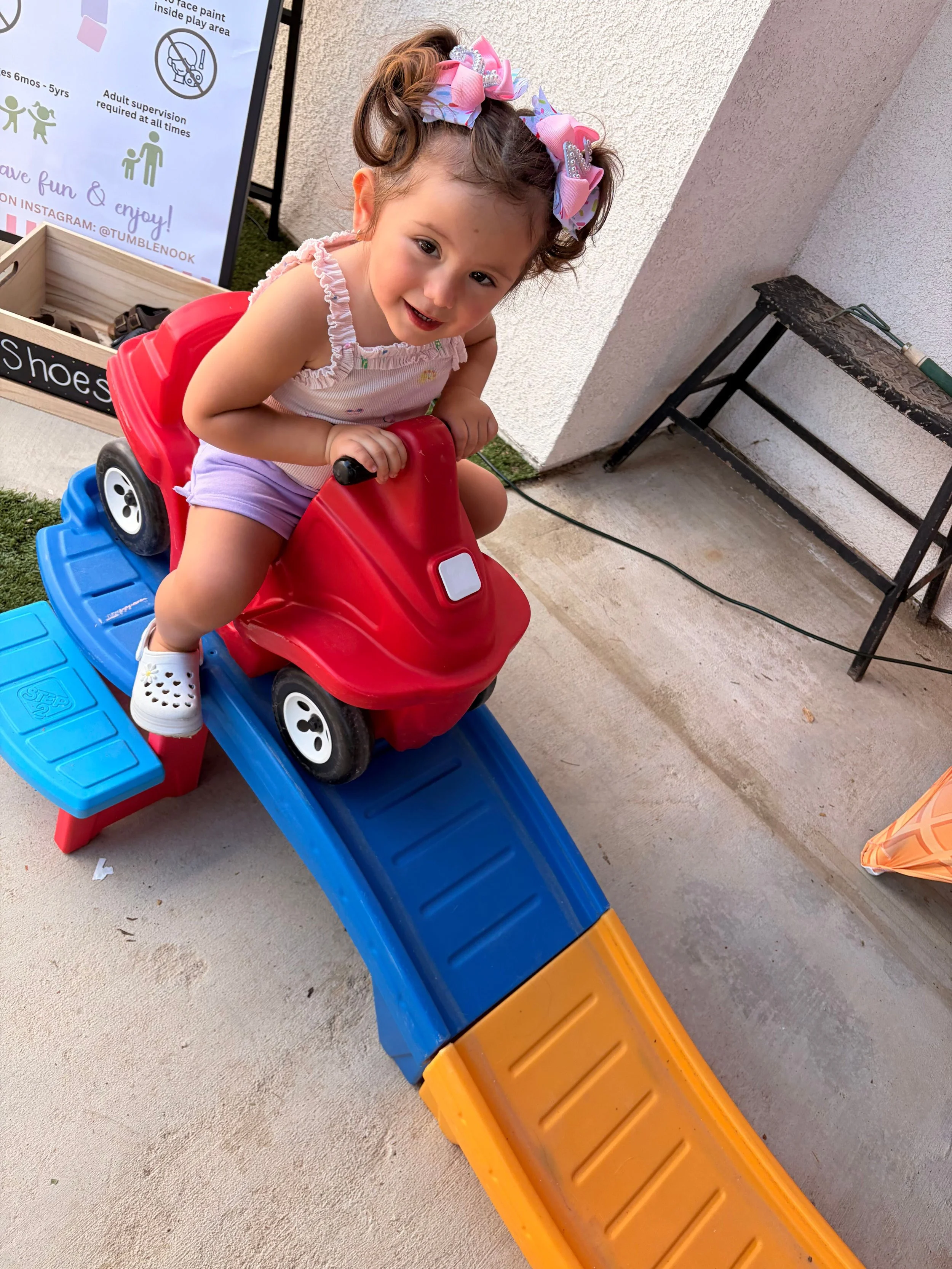 A young girl with curly hair tied with pink and purple bows, wearing a sleeveless top and shorts, is sitting on a plastic red ride-on toy car with a blue slide attached, outdoors on concrete ground near a textured wall and a black metal table.