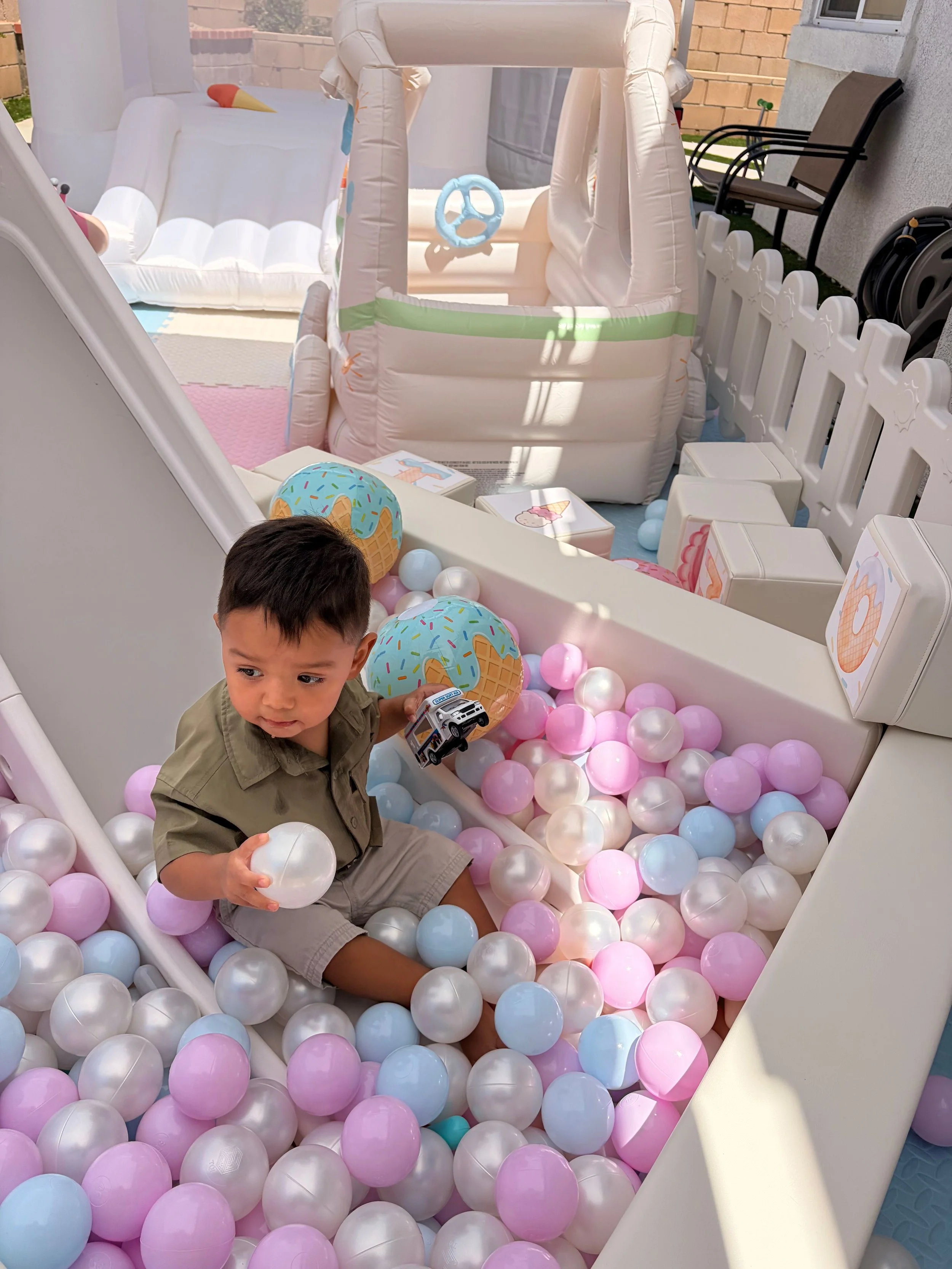 A young boy sitting in a ball pit filled with pastel-colored plastic balls, holding one ball and surrounded by other balls. Behind him, there is an inflatable play area with a small inflatable slide and a playhouse with a steering wheel. The setting 