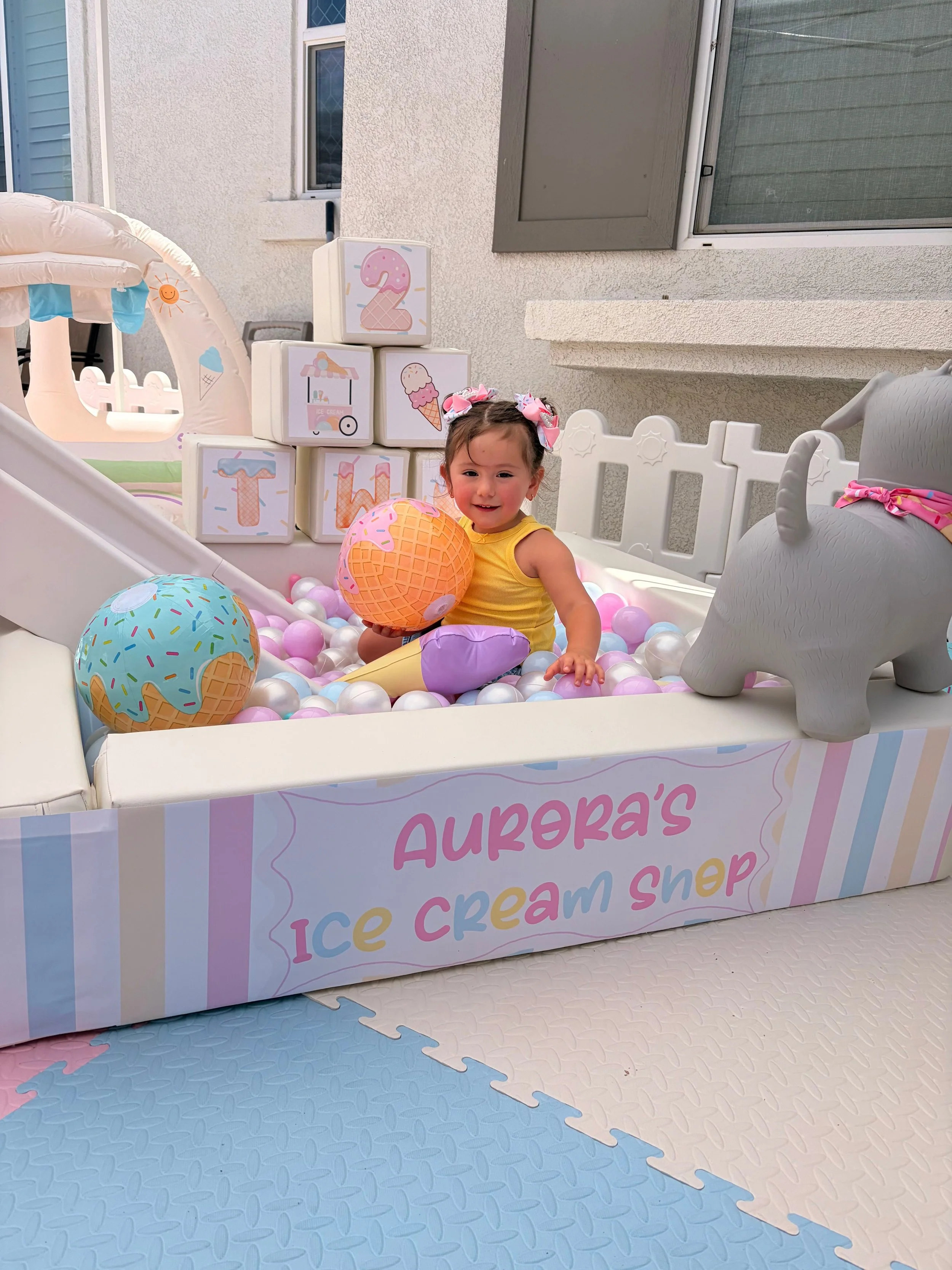 Young girl in a yellow outfit with pink bows in her hair sitting in a ball pit filled with pink, purple, and white balls, holding an ice cream cone-shaped ball. The scene is outdoors with a birthday-themed ice cream shop setup, including decorative b
