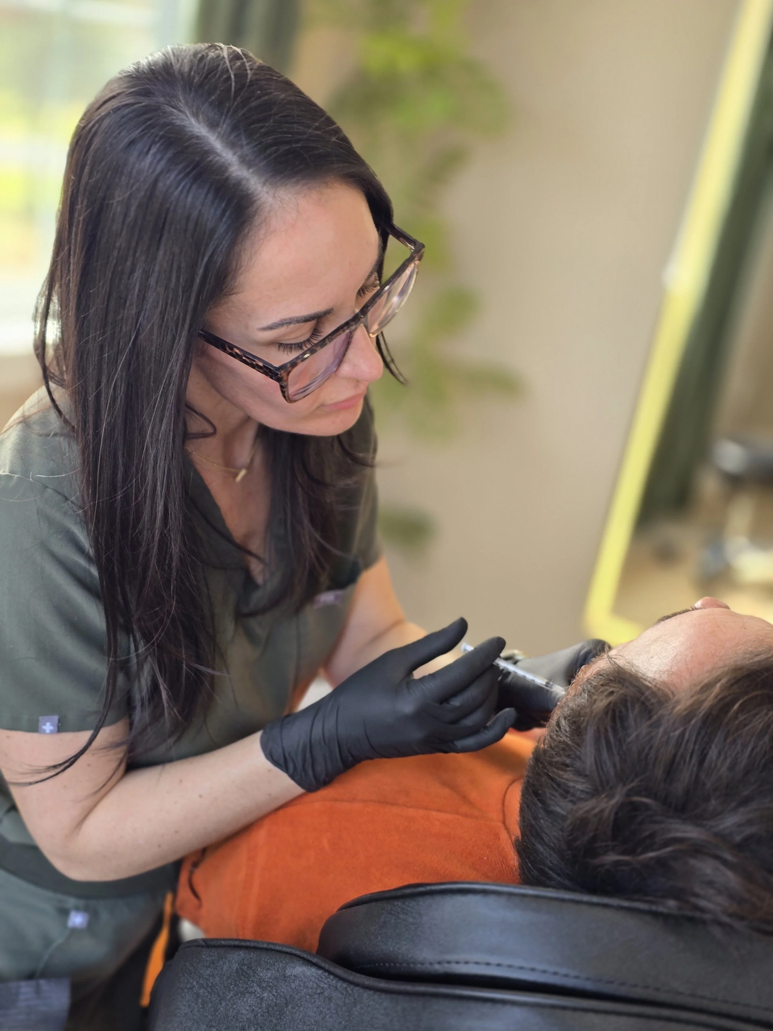 Tattoo artist in green uniform and leopard print glasses tattooing a person's neck area.