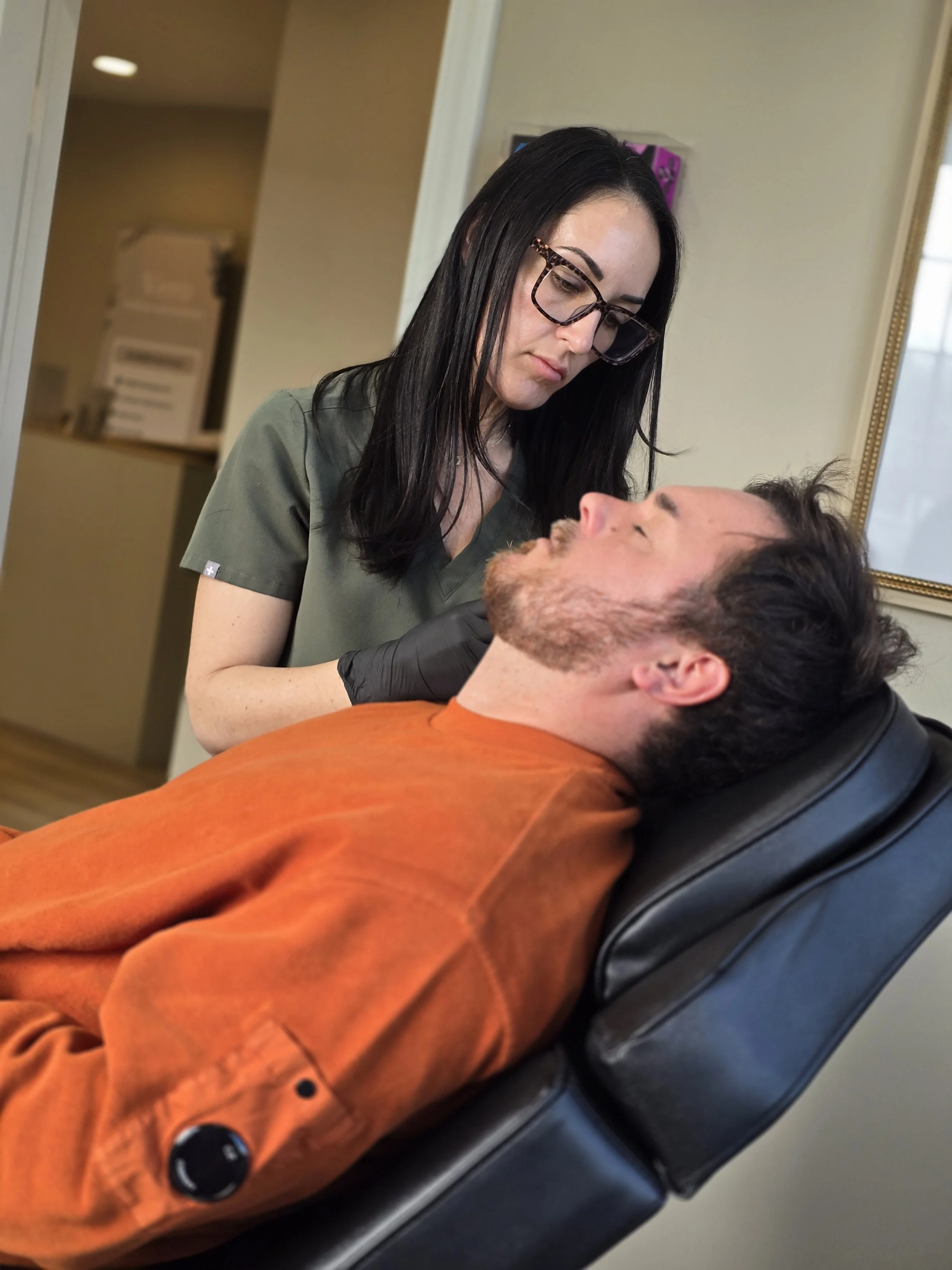 A woman with black hair, glasses, and medical scrubs is leaning over a man lying in a chair, possibly at a medical or dental office.