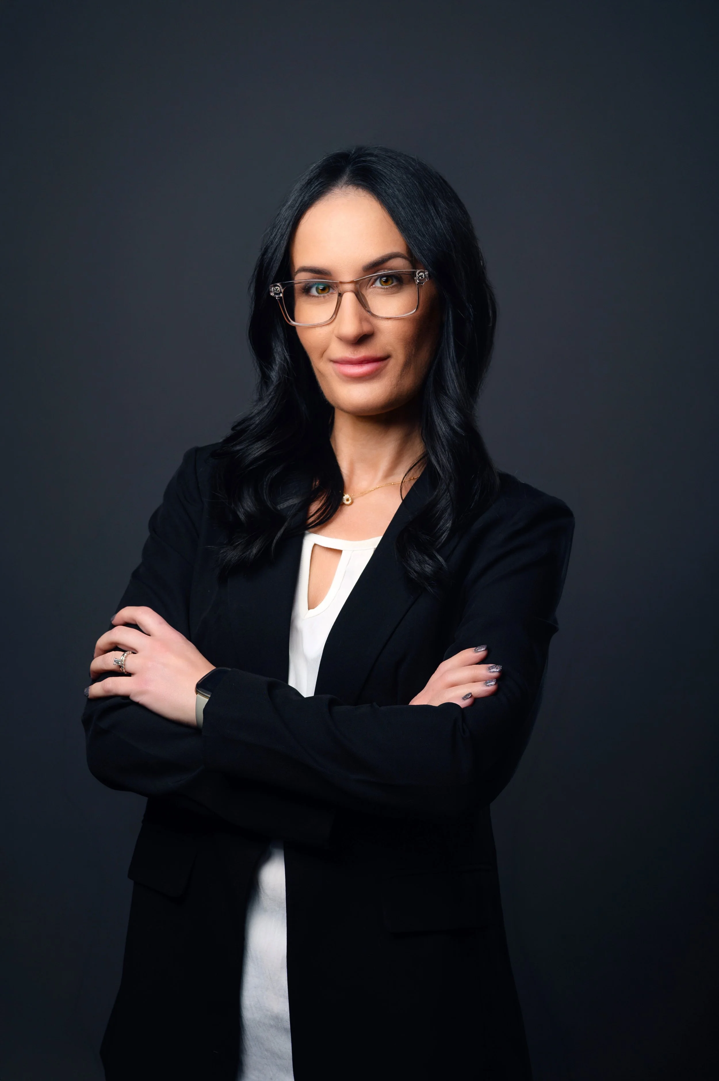Portrait of a confident woman with black hair, glasses, and professional attire standing with arms crossed against a dark background.