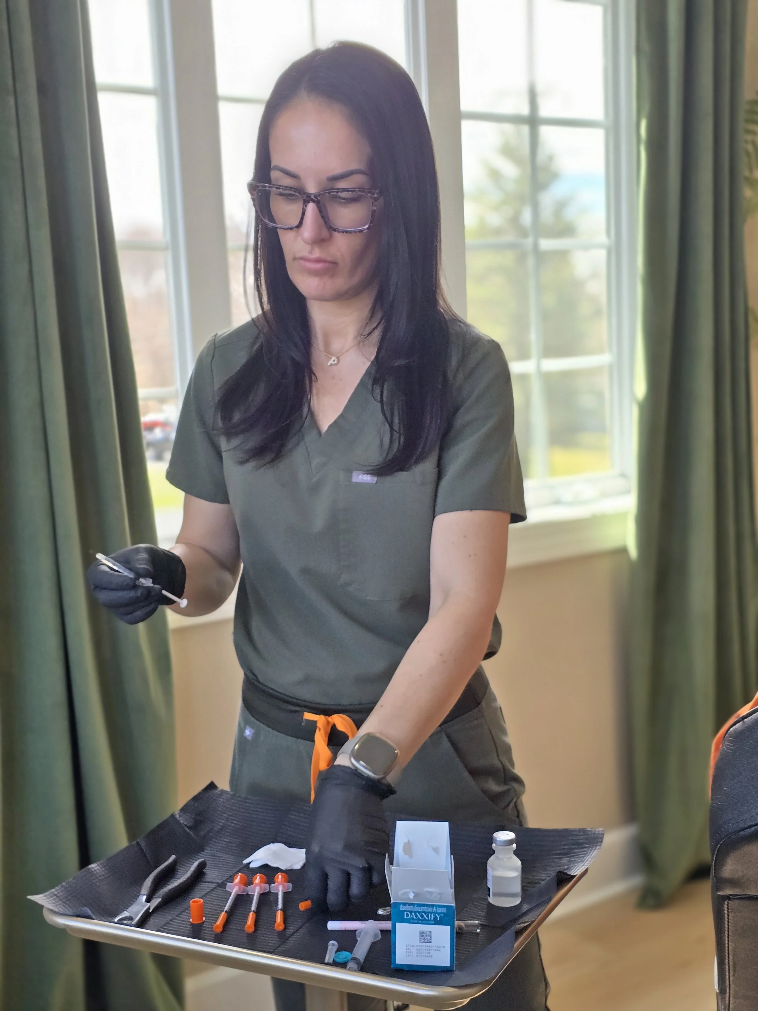 A woman medical professional with black hair, glasses, and scrubs is preparing syringes on a tray with vaccine supplies, including syringes, alcohol wipes, and bottles, in a clinical setting with sunlight coming through a window.