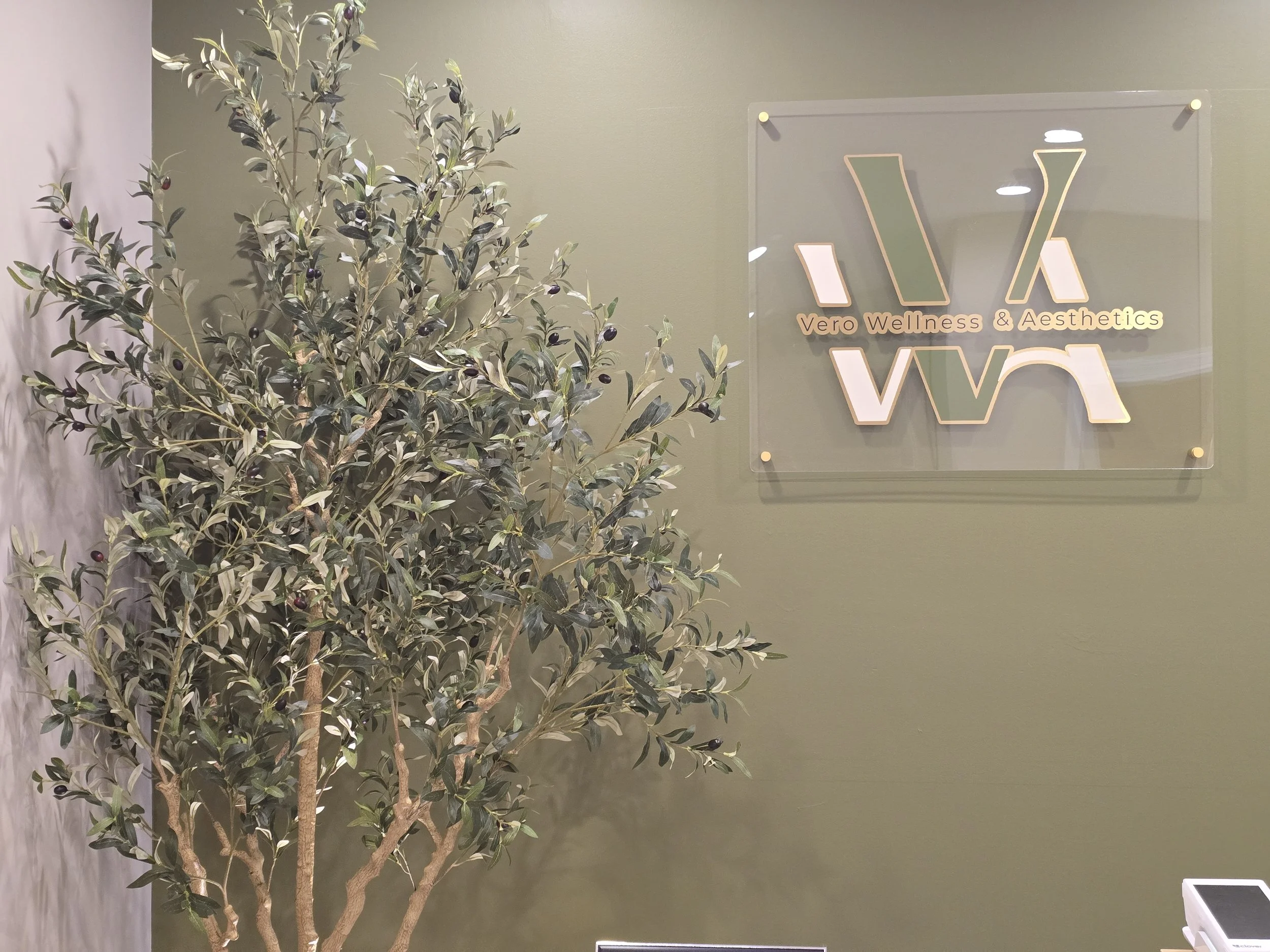 Interior of a wellness and aesthetics clinic with a green wall, an olive tree, and a sign that reads "Vero Wellness & Aesthetics" with a stylized "V" logo.