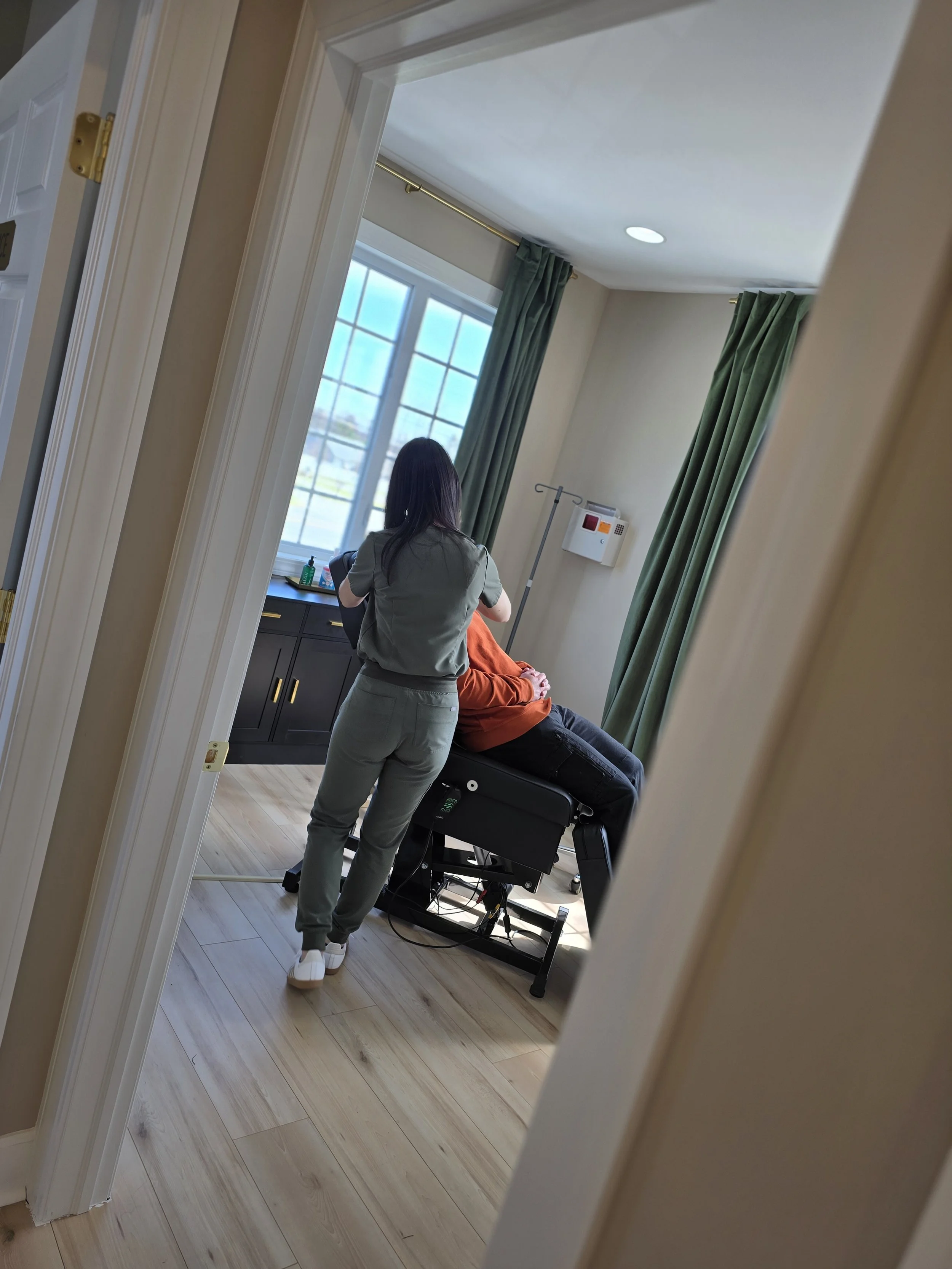 Reflection of a person in a mirror showing a healthcare worker preparing a patient for a medical procedure in a room with large windows and green curtains.