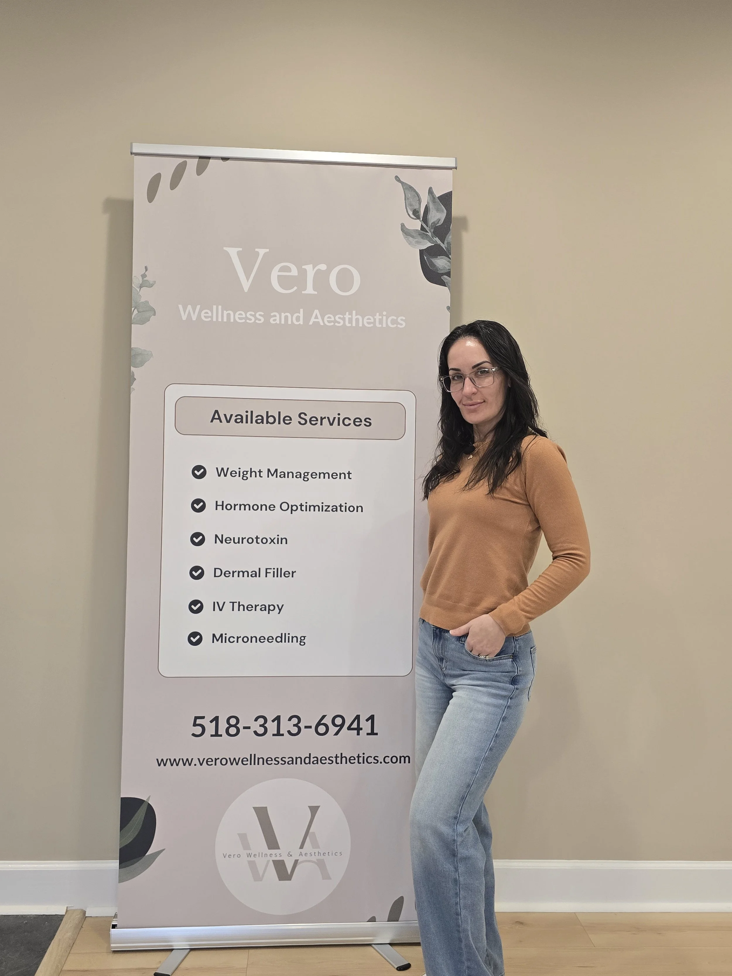 A woman standing next to a vertical banner for Vero Wellness and Aesthetics, which lists available services such as weight management, hormone optimization, neurotoxin, dermal filler, IV therapy, microneedling, and displays a phone number and website.