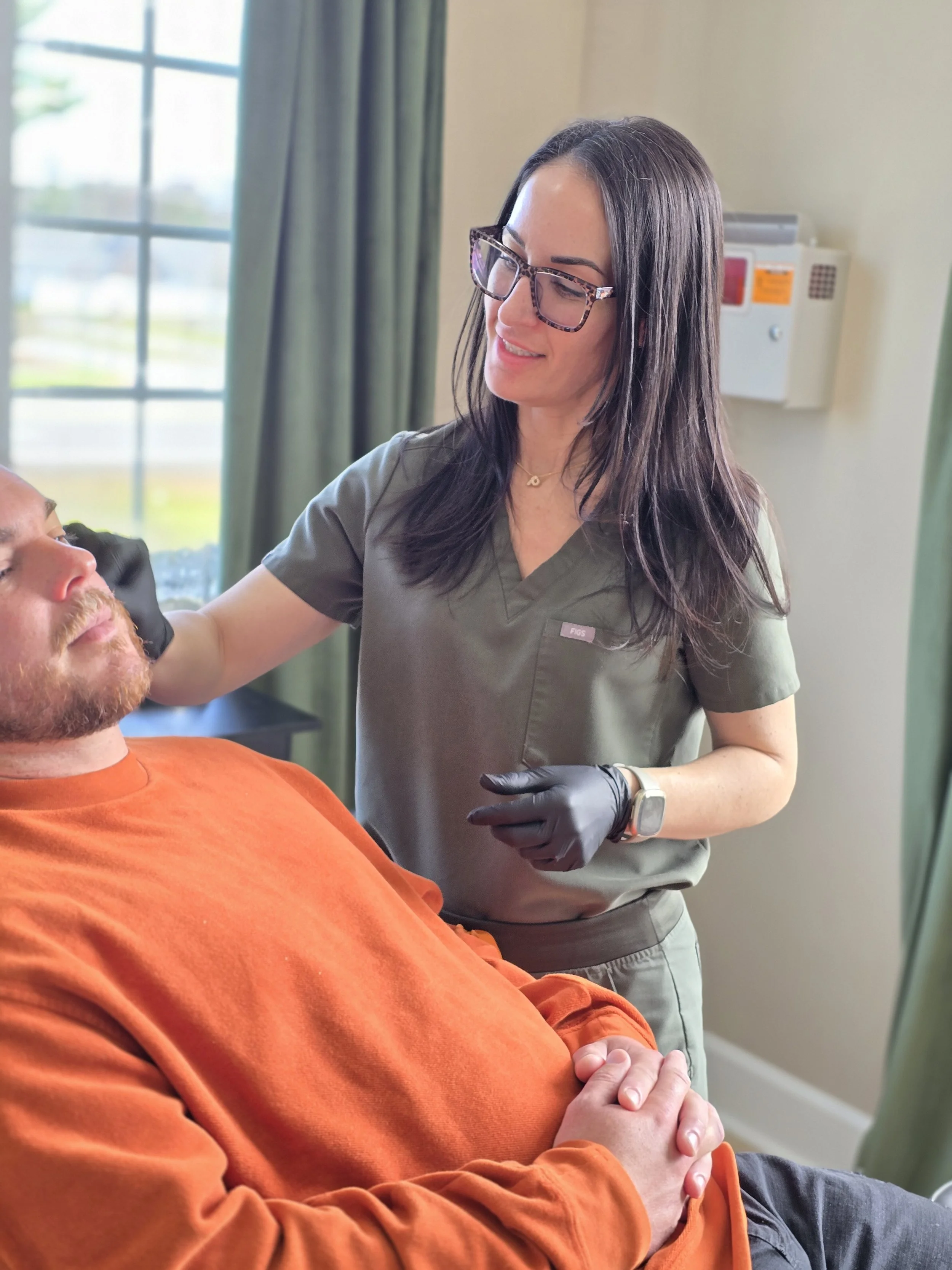 A female healthcare worker with long dark hair, glasses, and a gray uniform is smiling as she interacts with a man lying in a hospital bed. The man has a beard, is wearing an orange shirt, and has his hands clasped. The scene is set in a medical room with a window and green curtains in the background.