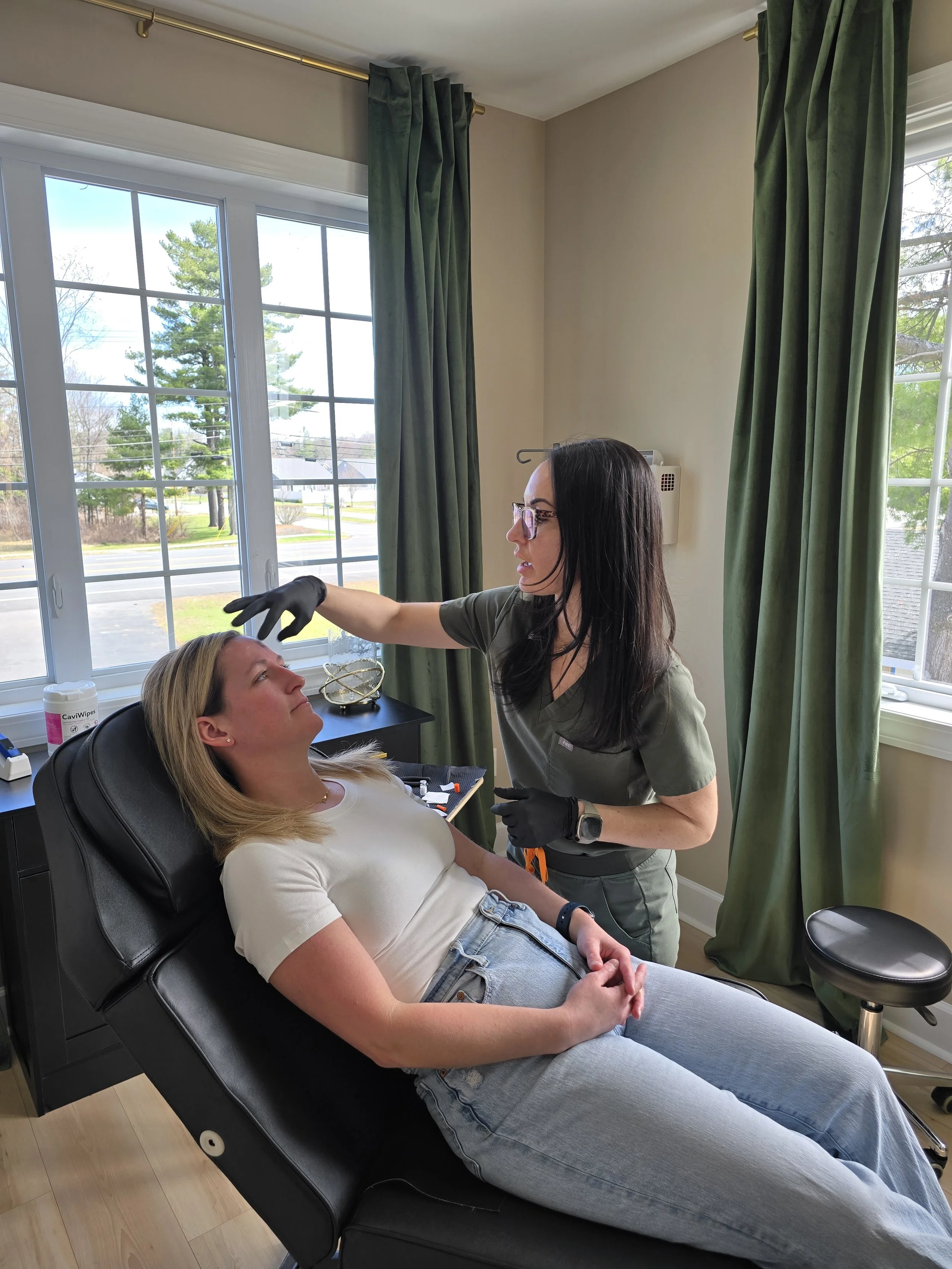A woman lying on a medical chair, receiving a cosmetic or medical procedure from a professional woman in a healthcare setting with large windows and green curtains.