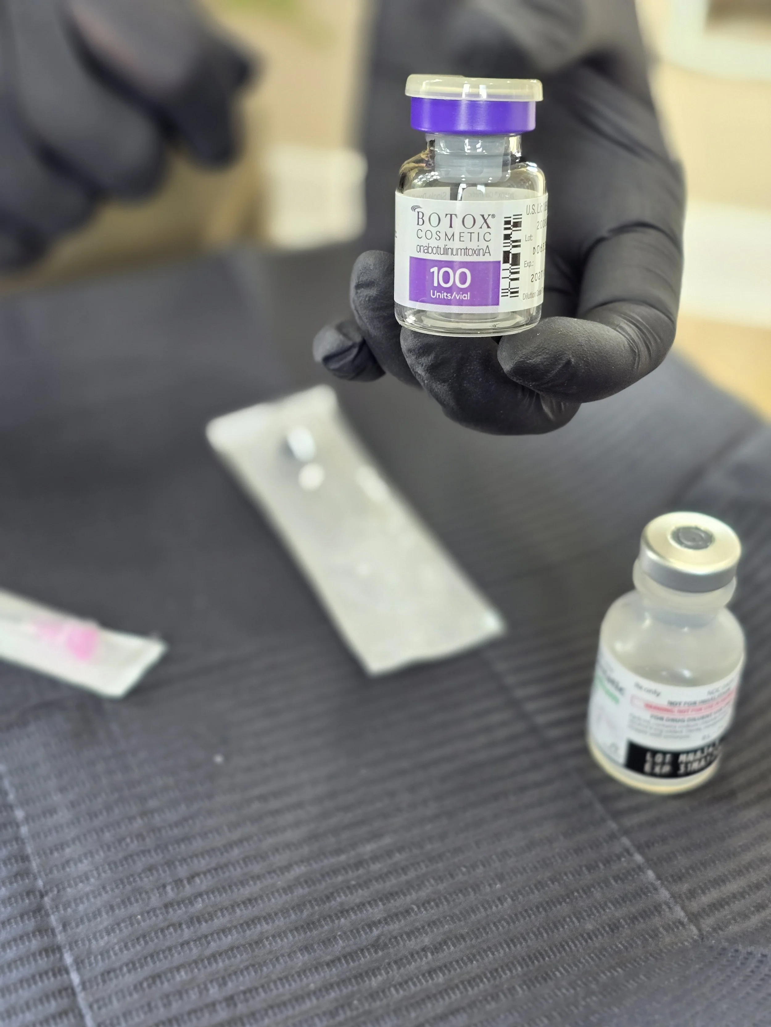 Medical vials, including Botox and another medication, on a dark textured surface with a syringe and small papers nearby.
