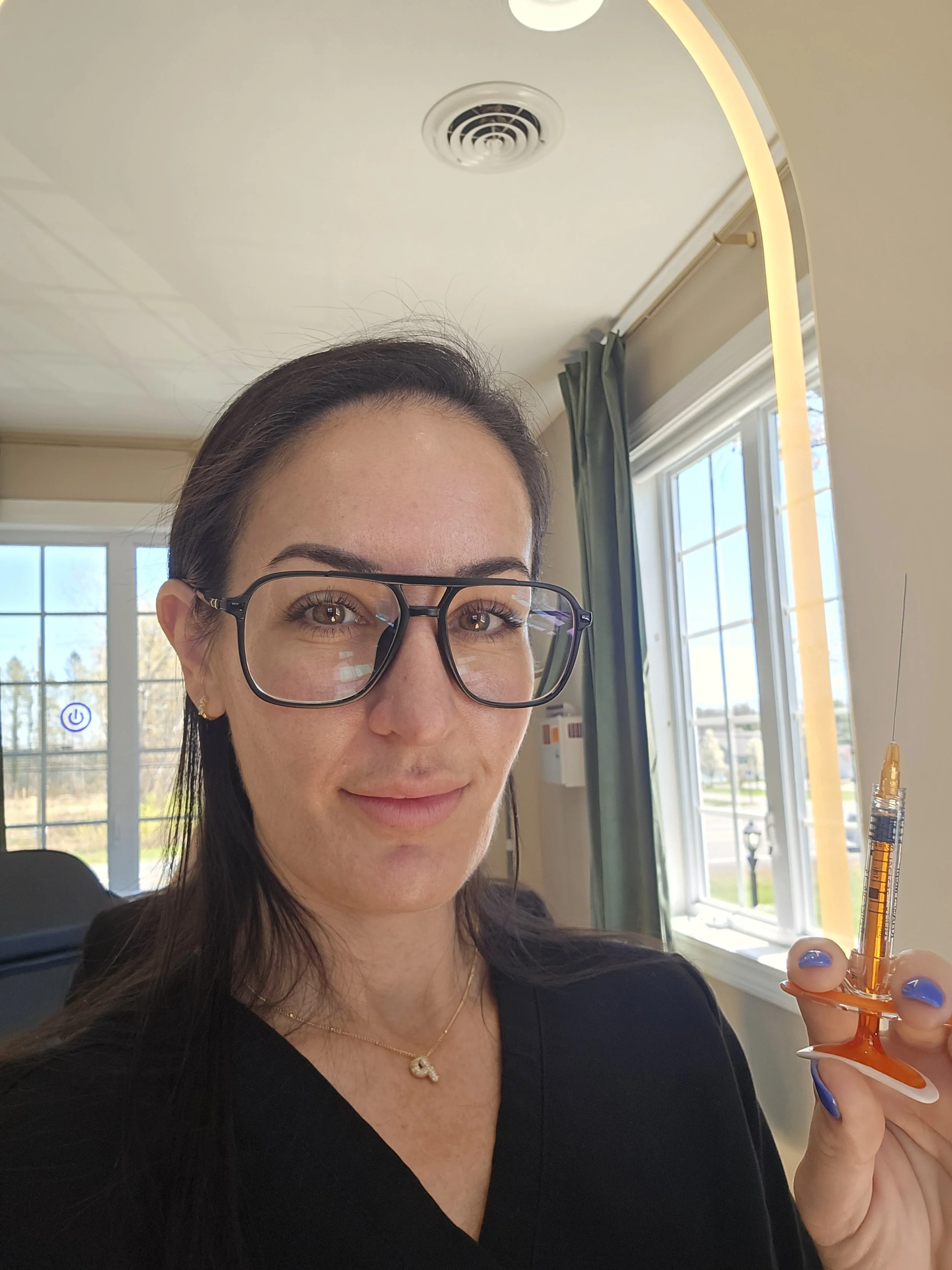 A woman wearing glasses and a black top, holding a syringe, standing near a window in a room with beige walls and curtains.