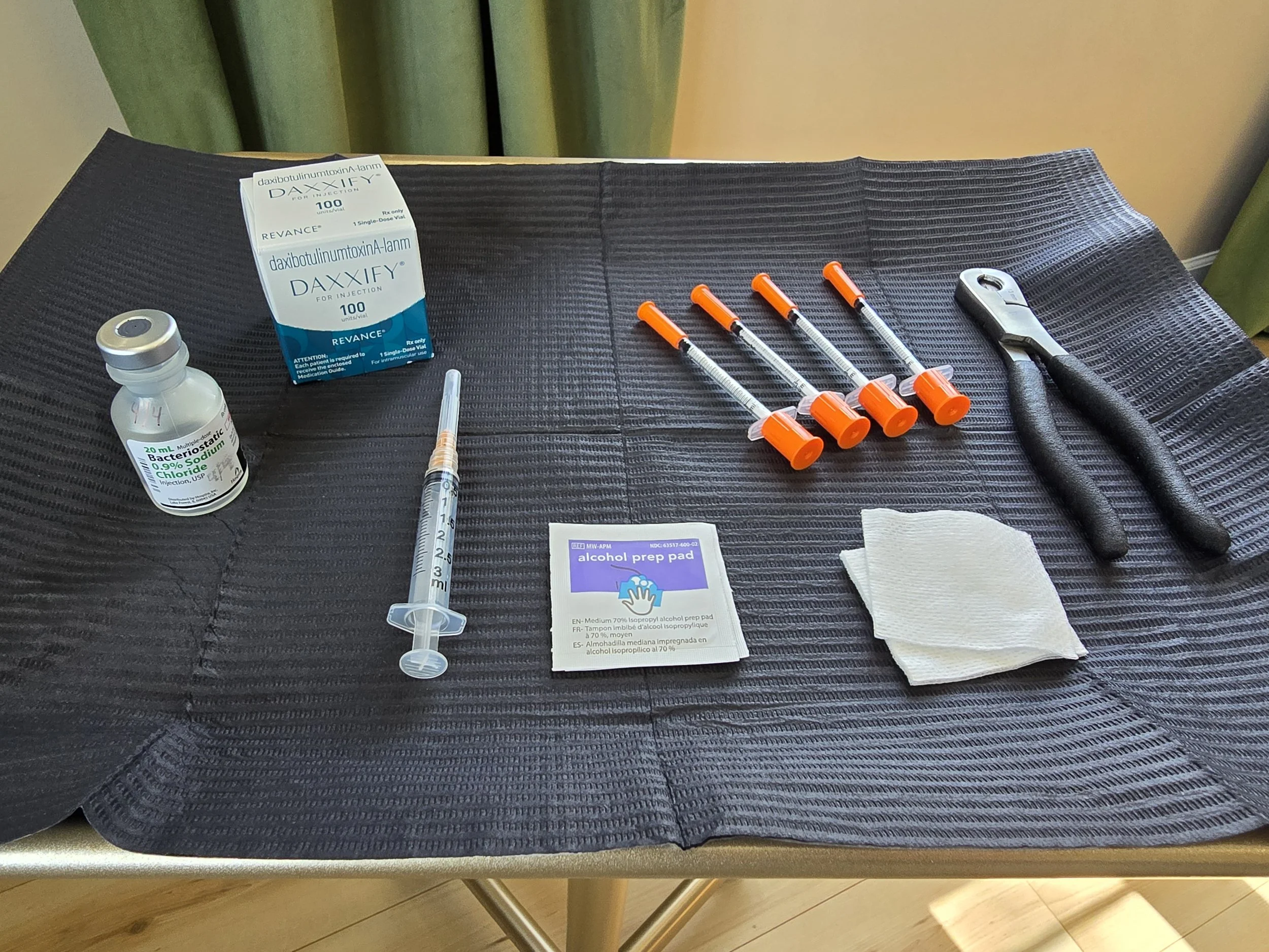 Medical supplies on a table including a box of DAXXIFY for injection, a vial of bottles, syringes, a puncture device, alcohol prep pad, and paper towels.