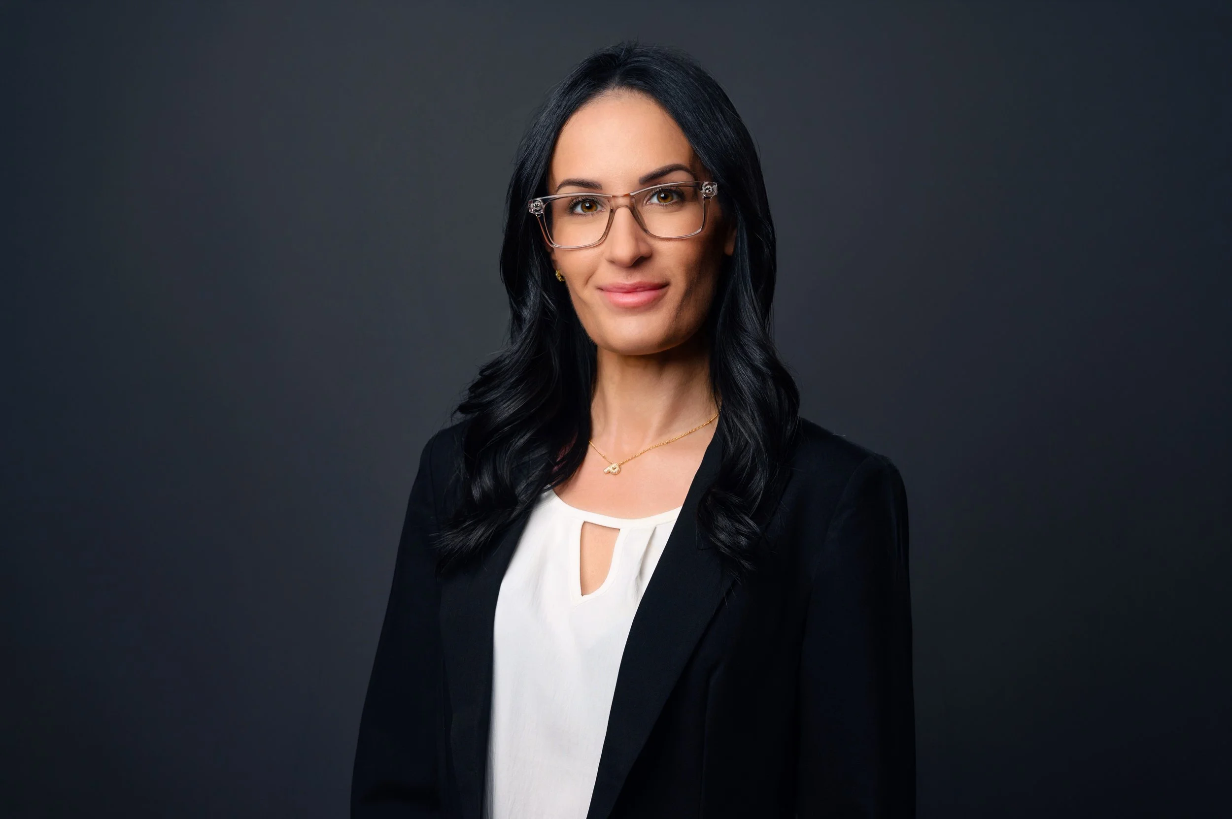 A woman with long black hair, wearing glasses, a necklace, and a professional outfit, posing against a dark gray background.