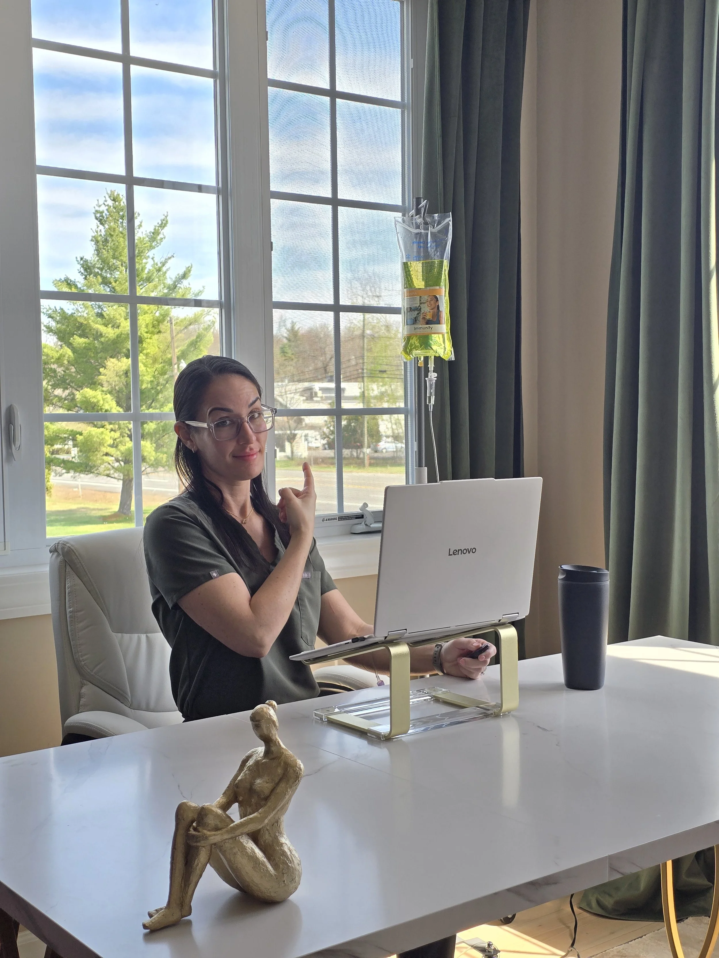 A woman with glasses and dark hair sitting at a white table with a Lenovo laptop in front of her. She is holding a computer mouse with her right hand and pointing upward with her left index finger. Behind her, there is an IV fluid bag hanging on a stand with a yellowish liquid inside. Outside the window, trees and a parking lot with cars are visible.