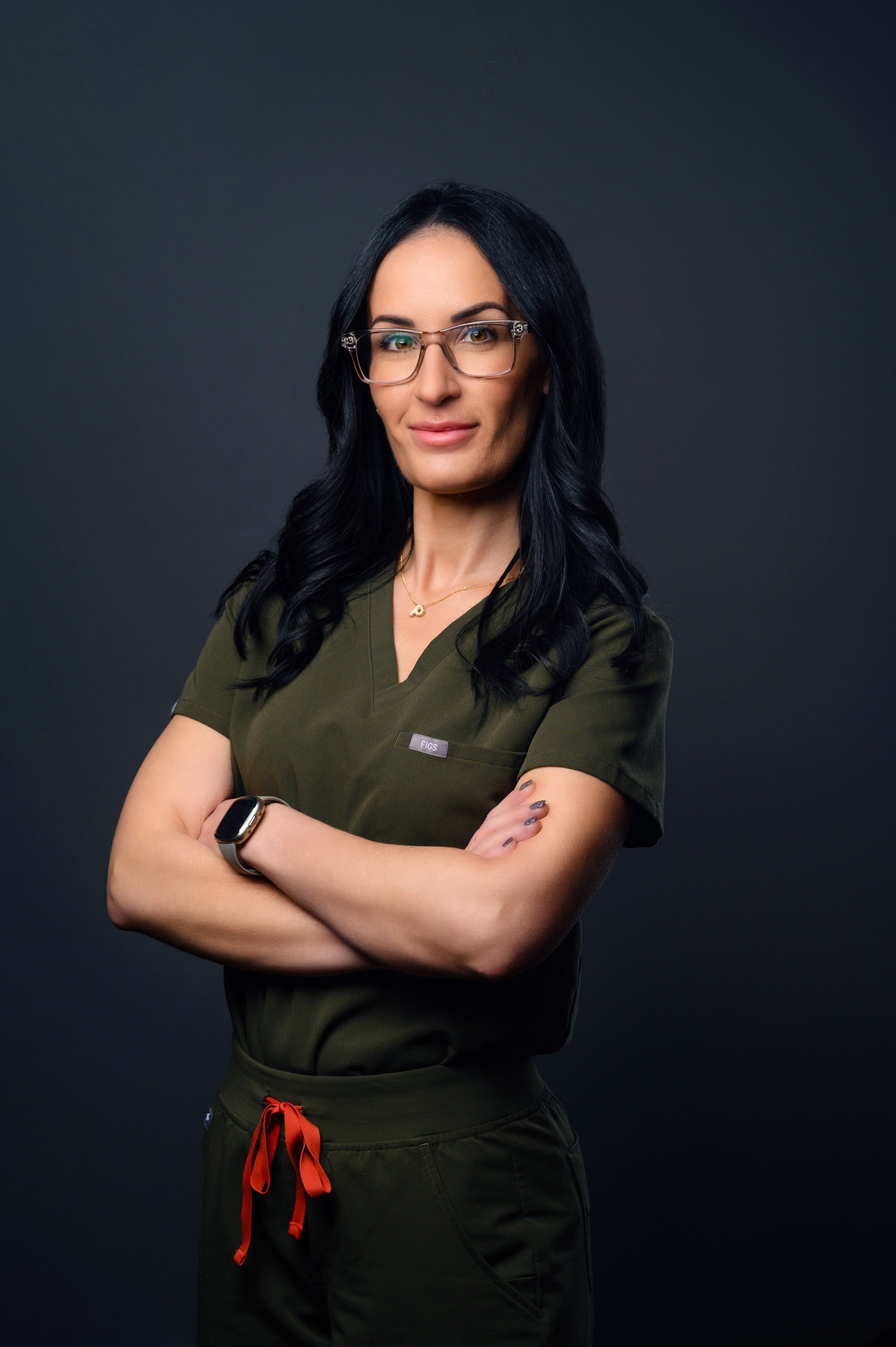 A woman with long black hair, glasses, and a confident expression, wearing a dark green scrub top with a small logo, a necklace, a smartwatch, and dark cargo pants with red drawstrings, stands with her arms crossed against a dark background.