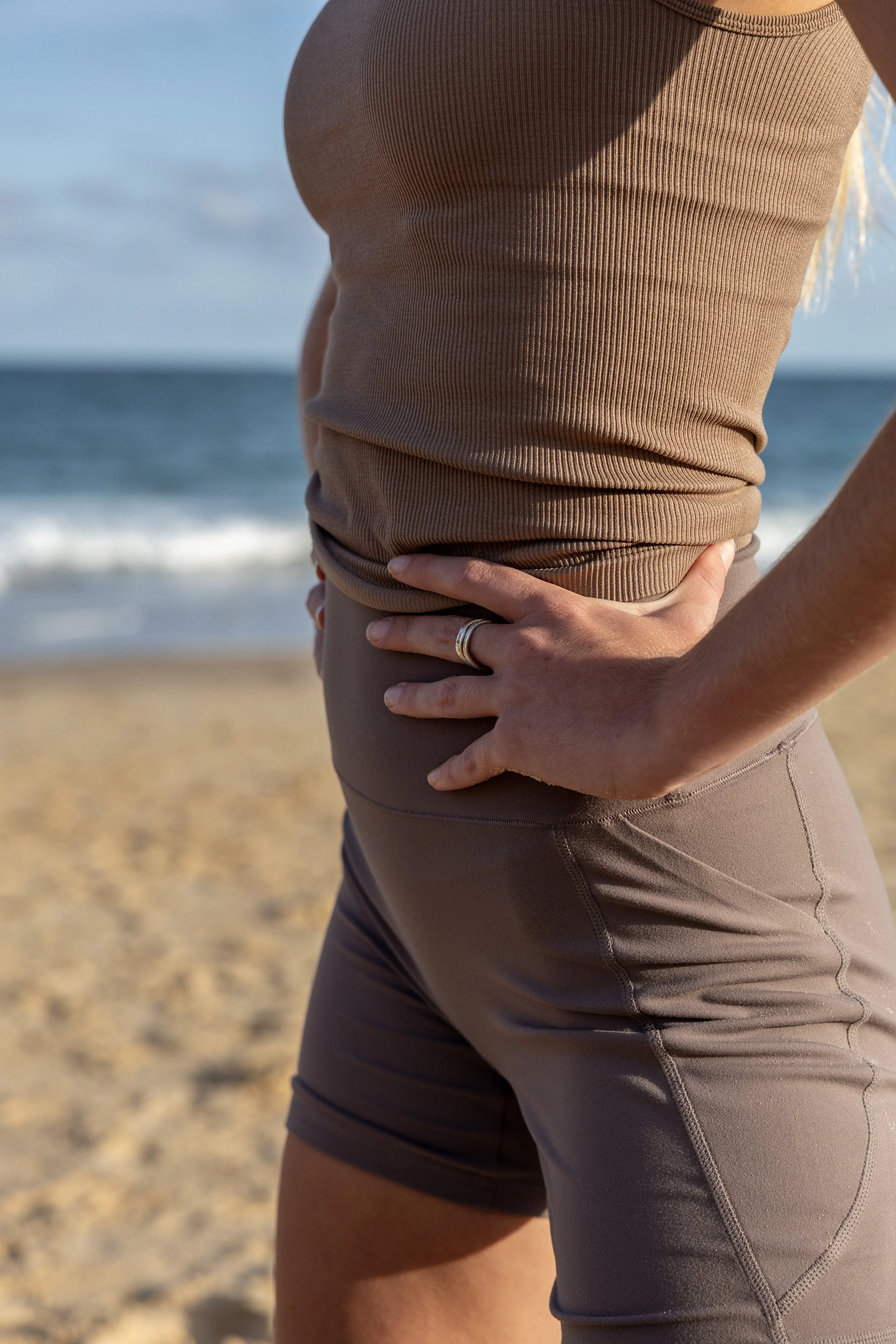 A person standing on the beach with one hand on their hip, wearing a beige or tan sleeveless top and dark workout shorts.