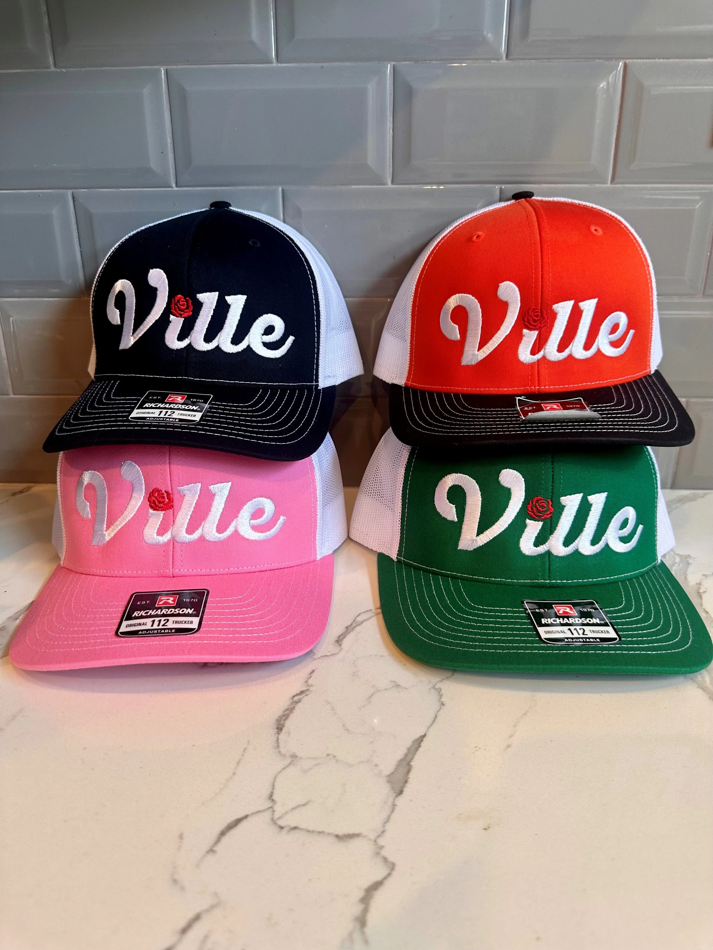 Four caps in black, pink, red, and green with the word 'Vile' embroidered on the front, featuring a small red rose above the 'i', displayed on a white marble surface with a gray tile background.
