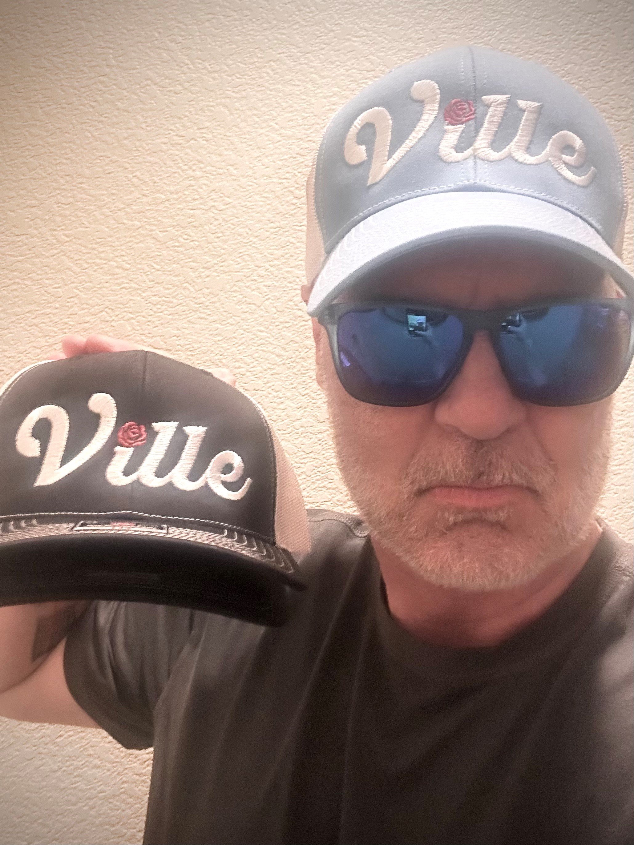 A man wearing a gray baseball cap with the word 'Viu' embroidered on it, dark sunglasses, and a black leather jacket, holding a black cap with the same 'Viu' logo in front of a beige textured wall.