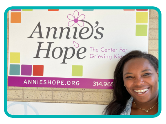 A Black woman with long dark hair smiling in front of a sign that reads 'Annie's Hope: The Center for Grieving Kids' with colorful squares and the website annieshope.org.