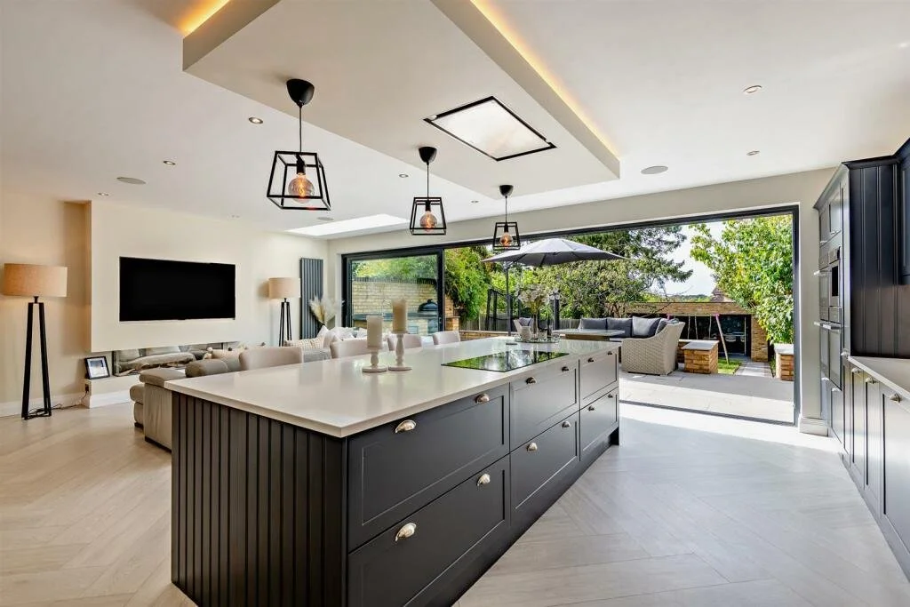 Modern open-concept kitchen and living room with black kitchen island, pendant lights, and large sliding glass doors leading to outdoor seating area with trees.