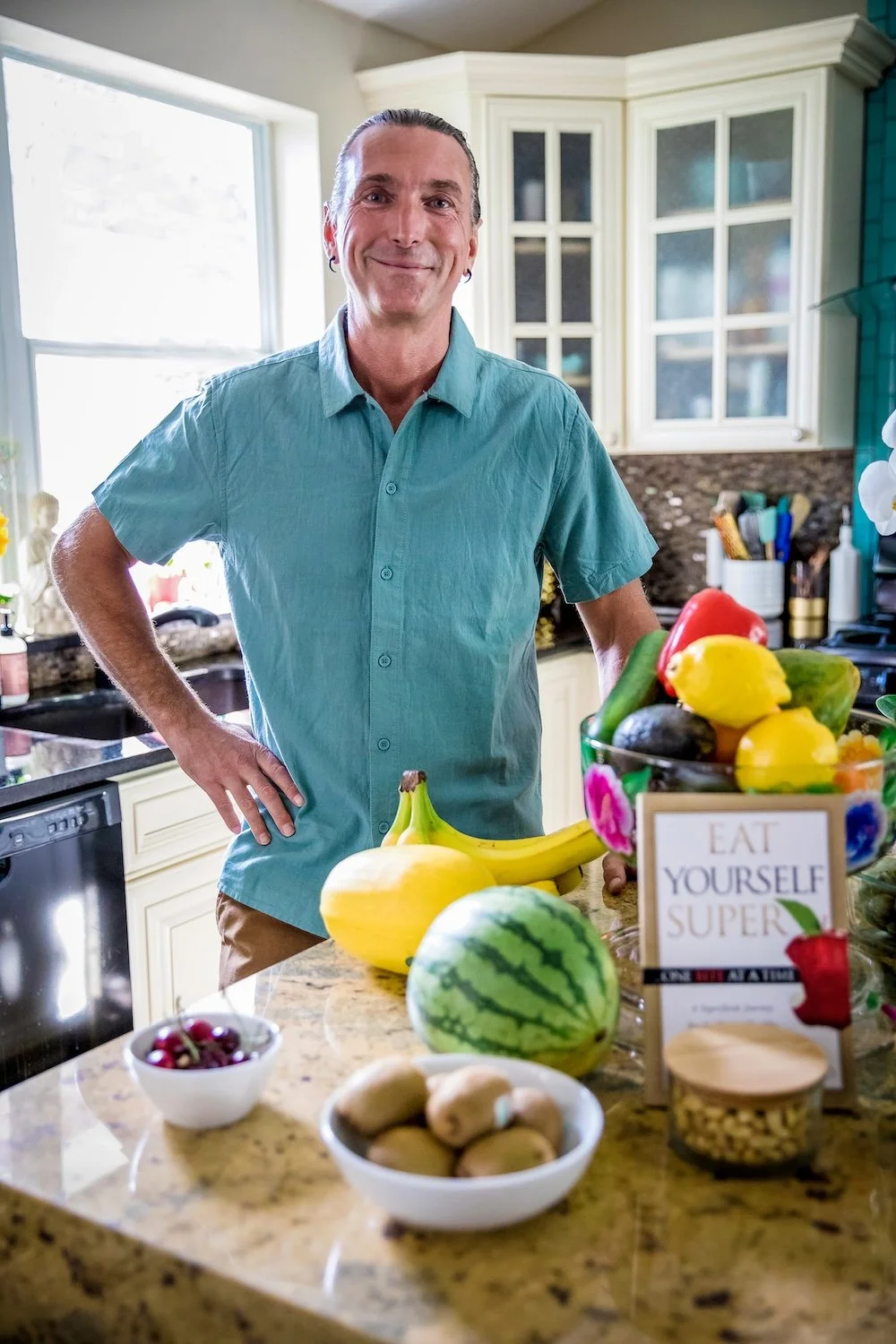 A man with gray hair and a blue button-up shirt stands in a kitchen next to a countertop with fresh fruits including bananas, a watermelon, kiwis, and a bowl of cherries. The background shows white cabinets and a window with sunlight coming through.