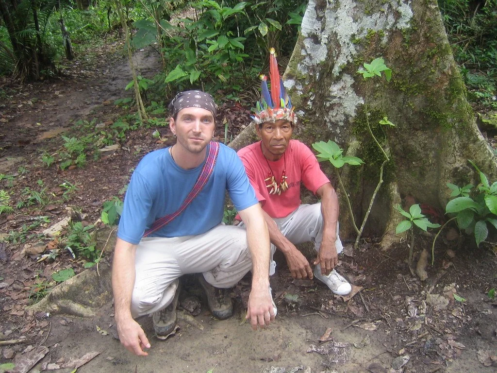 Two men squatting in a jungle, one wearing a blue t-shirt and bandana, the other wearing a red t-shirt and a colorful headdress.