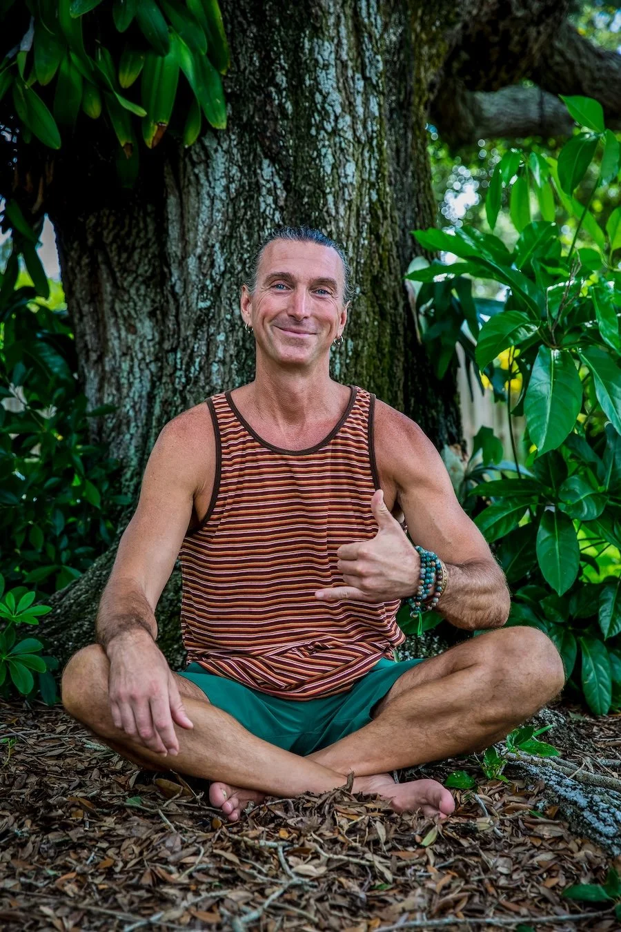 A man sitting cross-legged on the ground in front of a large tree, surrounded by green leaves, smiling, wearing a striped tank top and teal shorts, making a shaka sign with his right hand.
