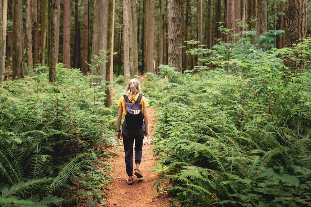 A woman with blonde hair walking on a narrow dirt trail through a green forest with tall trees and dense foliage.
