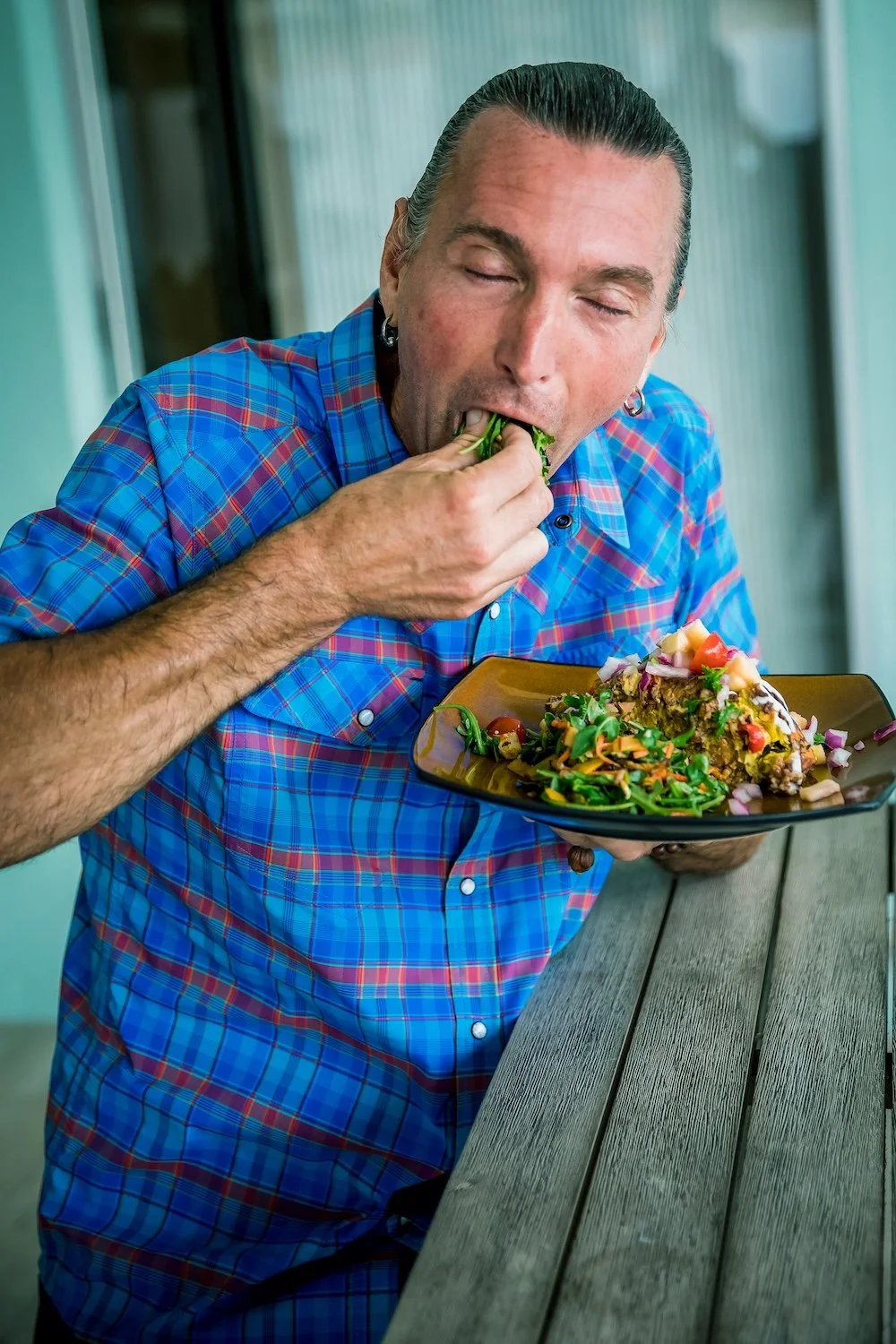 A man wearing a blue plaid shirt is eating a salad with vegetables and possibly a taco or burrito, with plants and a patio or balcony environment in the background.