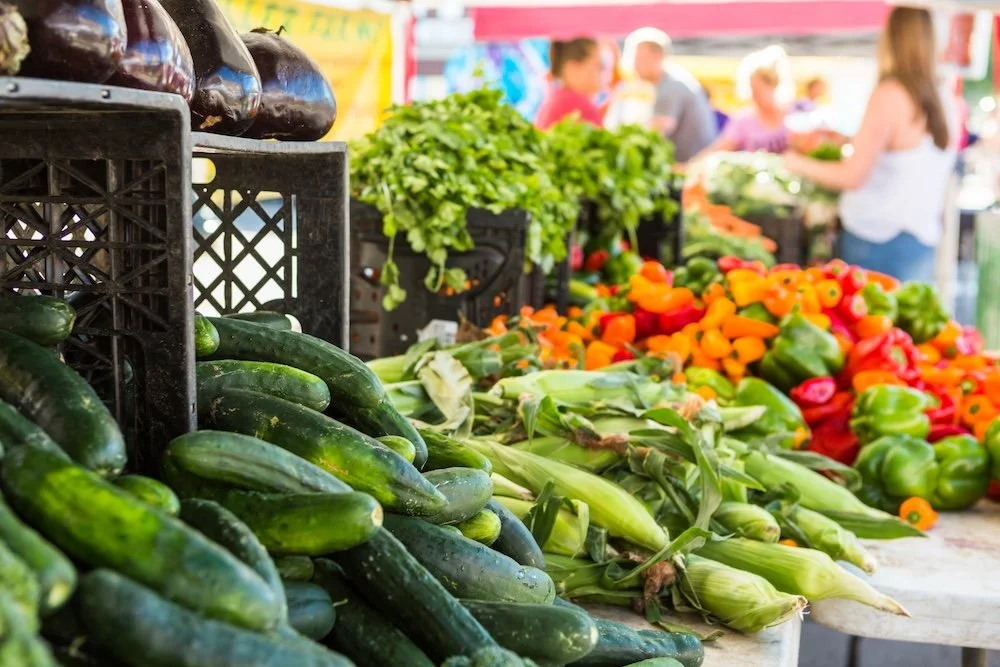 Fresh cucumbers, corn, and colorful bell peppers at a farmers' market stall with people shopping in the background.