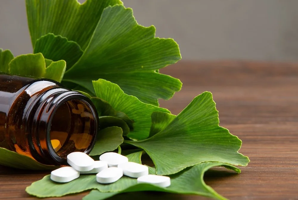 An open brown glass bottle lying on its side on a wooden surface, with white pills spilled out onto green ginkgo leaves surrounding it.