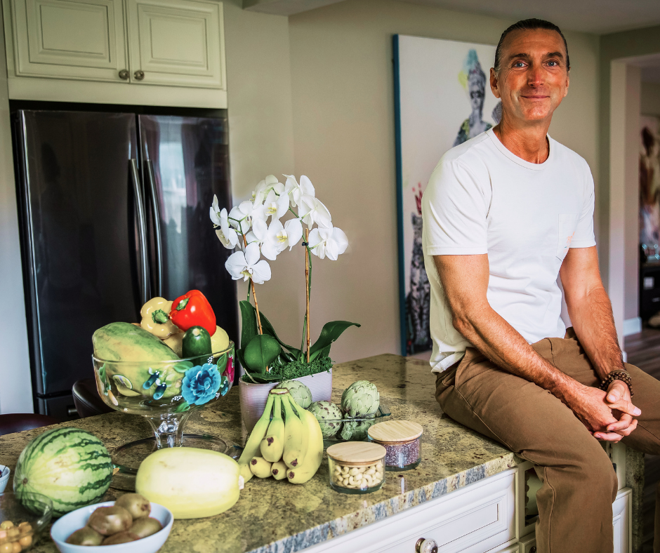 A middle-aged man in a white t-shirt and khaki pants sitting on a kitchen counter next to a bowl of fruit, potted white orchids, and various vegetables and fruits. The kitchen has a modern look with light-colored cabinets and a black refrigerator.