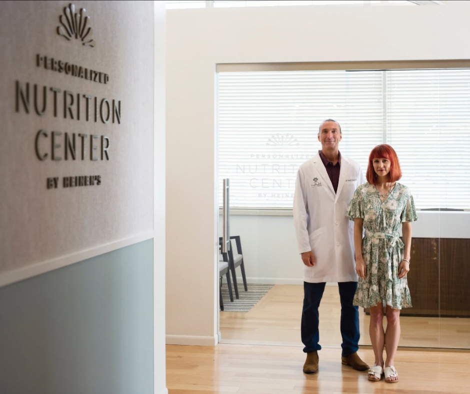 A man in a white medical coat and a woman in a floral dress standing inside a nutrition center with a sign on the wall and a large window with blinds in the background.