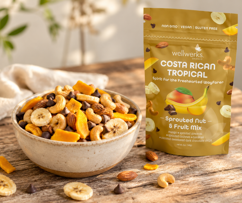 A bowl of mixed trail mix with nuts and dried banana slices next to a yellow package of Wellwerks Costa Rican Tropical sprouted nut and fruit mix on a wooden table.