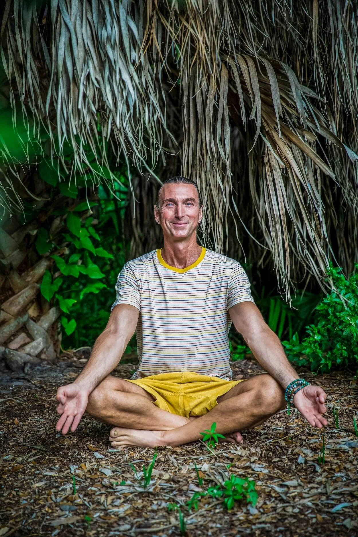 A man sitting cross-legged on the ground in a forest, smiling, with his hands resting on his knees in a meditative pose.