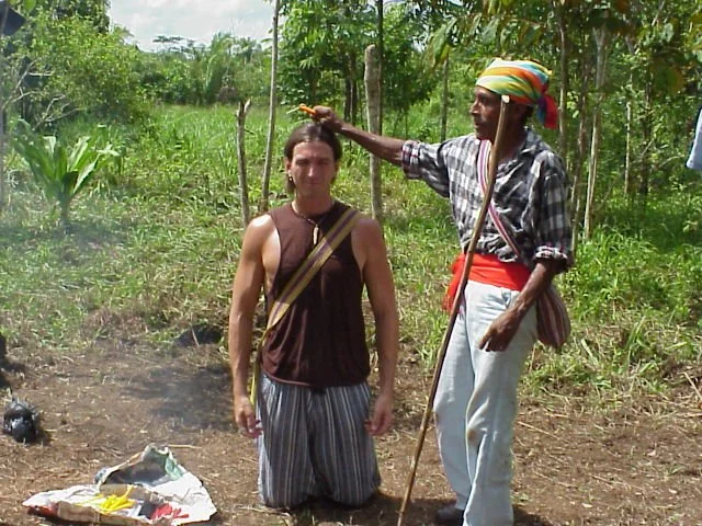A man kneels on the ground with his hands at his sides, while another man stands beside him, pointing at his head with a stick. The scene is set outdoors in a green, tropical environment.
