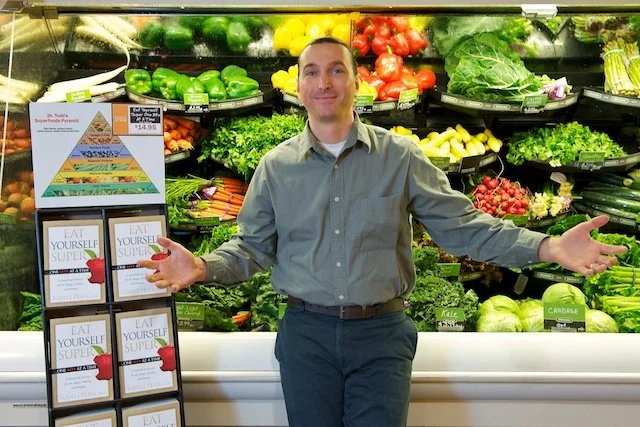 Man standing in front of a produce section at a grocery store, with fresh vegetables like peppers, lettuce, and tomatoes displayed behind him, and a book stand on the side.
