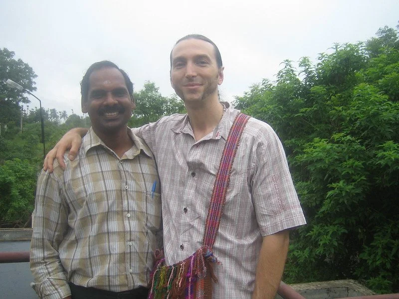 Two men standing outside near greenery, smiling. One man has dark hair and darker skin, wearing a plaid shirt. The other man has light hair and light skin, wearing a plaid shirt with a colorful shoulder bag. They have their arms around each other.