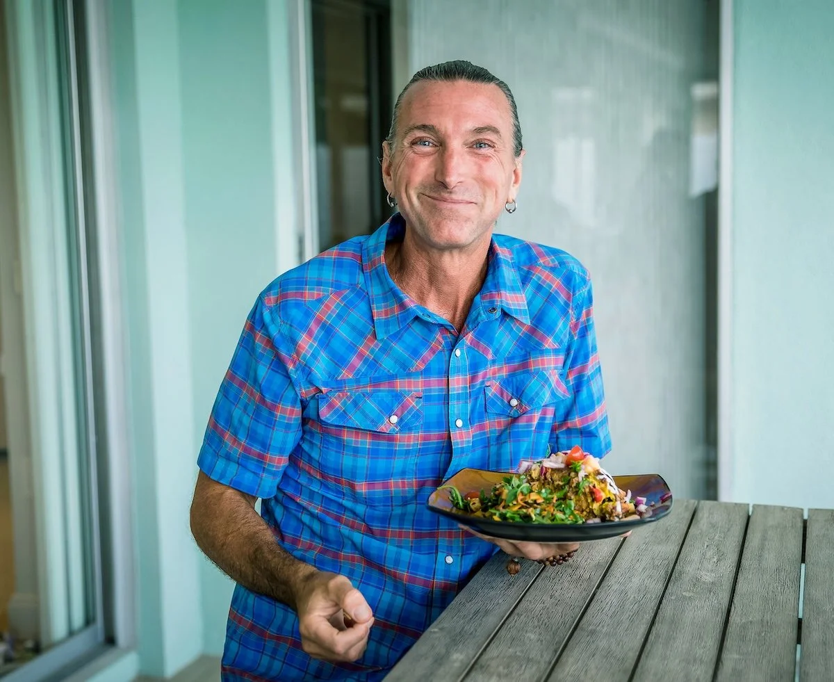 A smiling man with earrings holds a black plate of food, standing outdoors near a balcony or patio with a wooden table in front of him.