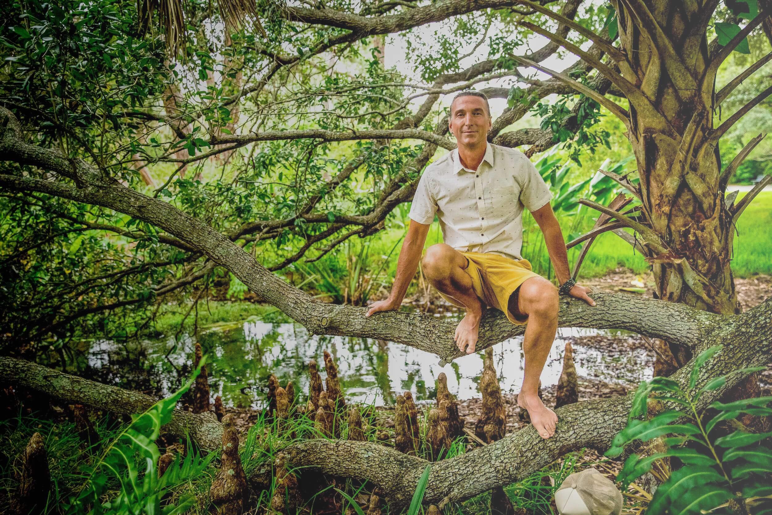 A man is sitting on a tree branch in a lush, green outdoor setting, surrounded by trees, water, and grass.