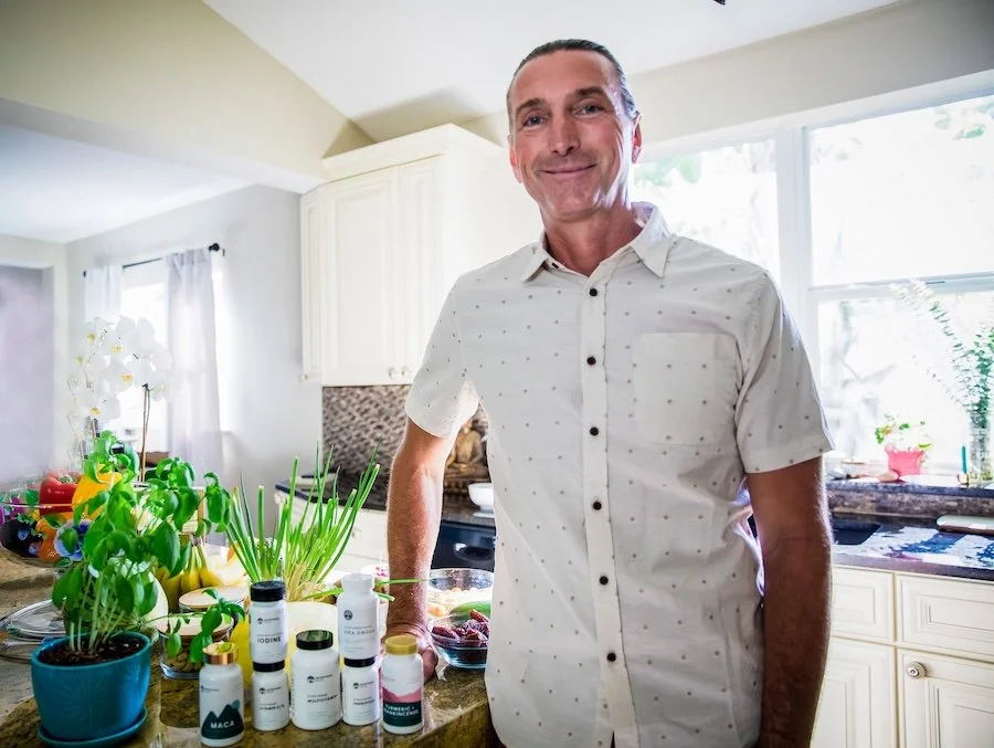 A man smiling in a bright kitchen, standing at a counter with bottles of dietary supplements and potted plants.