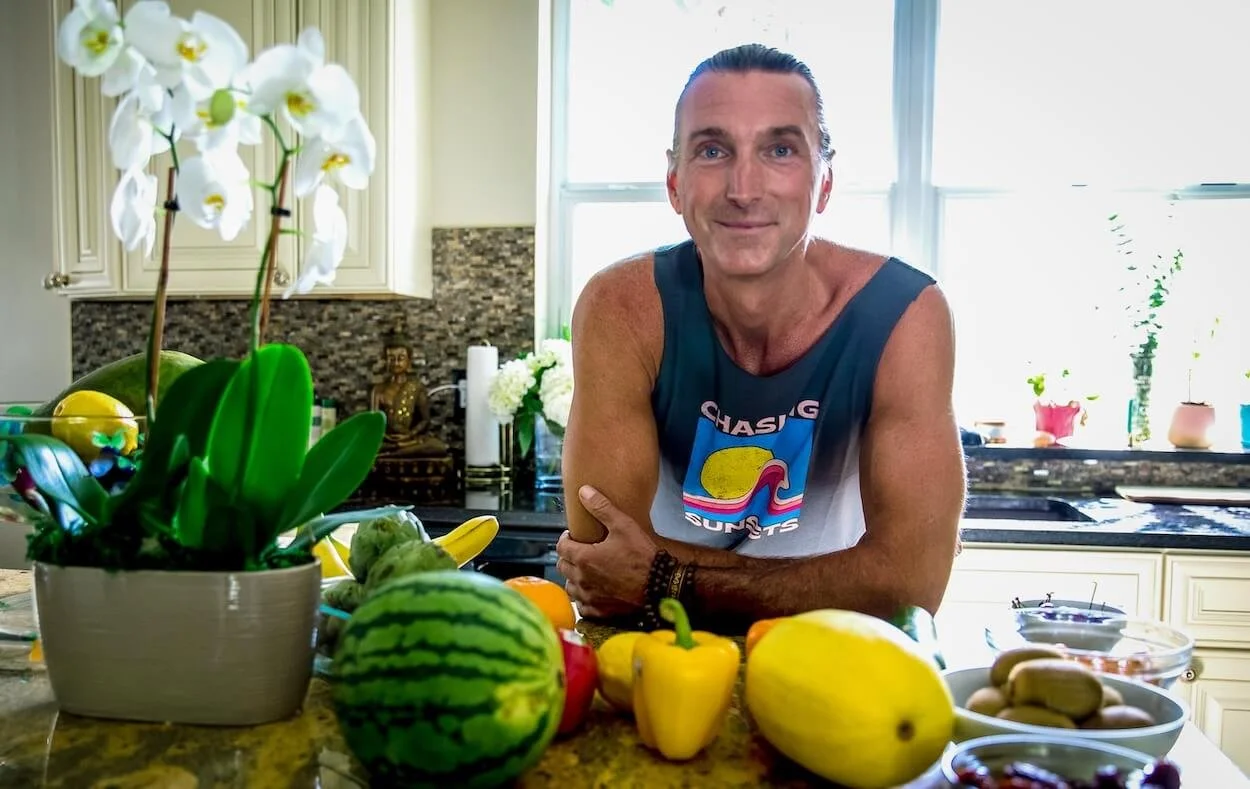 A man with a sleeveless shirt, smiling, in a bright kitchen with various fruits, vegetables, and a potted orchid on the counter.